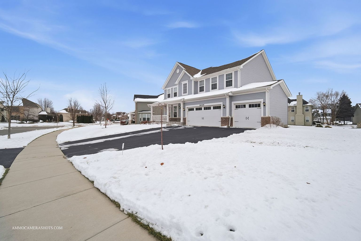 This is a front exterior view of a two-story house with light gray siding and white trim. The house features a two-car garage, a well-maintained lawn covered in snow, and a paved driveway. The sky is clear and blue, suggesting a bright winter day.