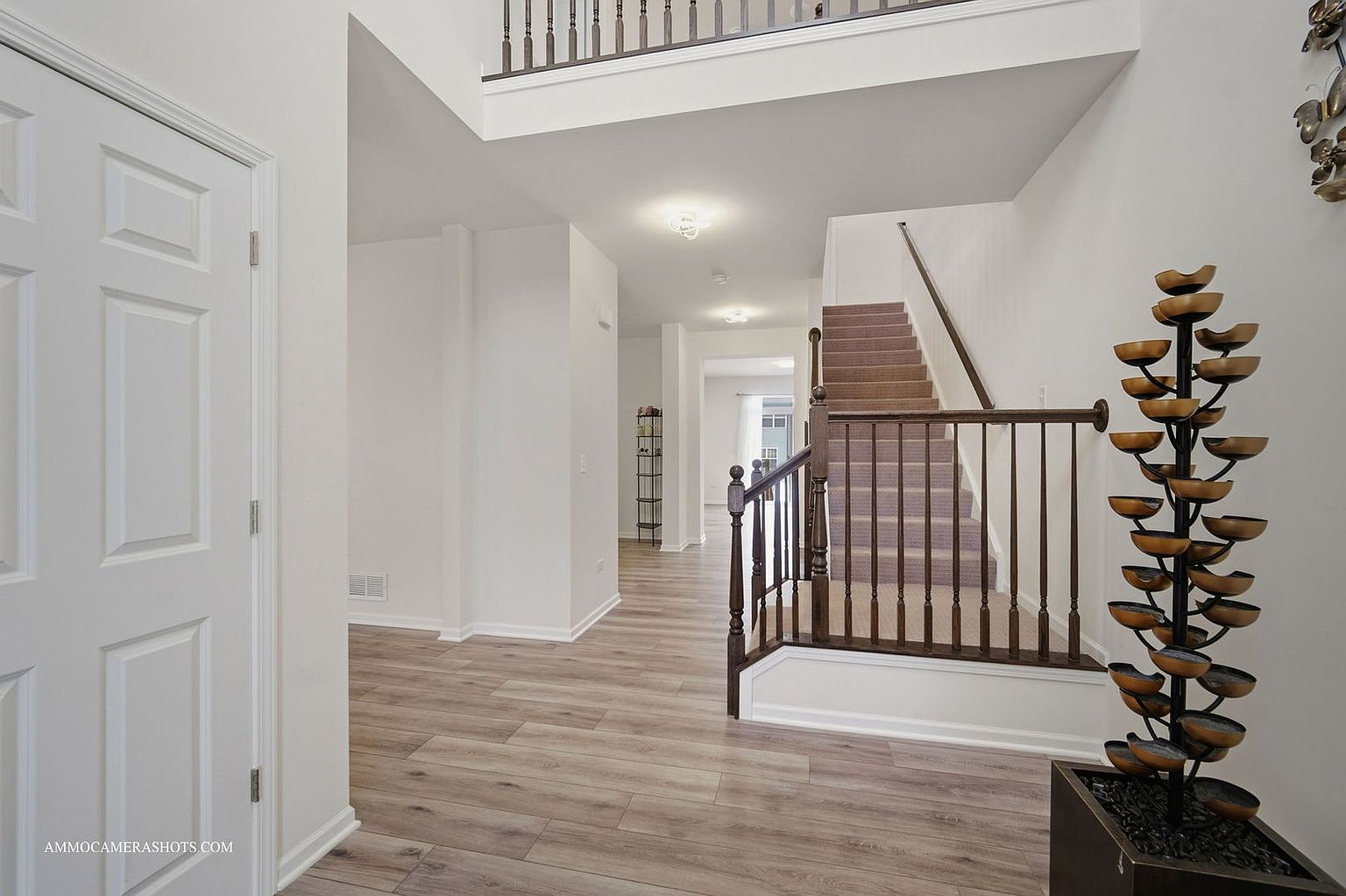 This interior shot showcases a bright and spacious hallway with a staircase leading to the upper level. The flooring is light-colored wood, and the walls are painted in a neutral tone, creating a clean and modern aesthetic. A decorative water fountain adds a unique touch to the space, while the staircase features dark wood railings and carpeted steps.