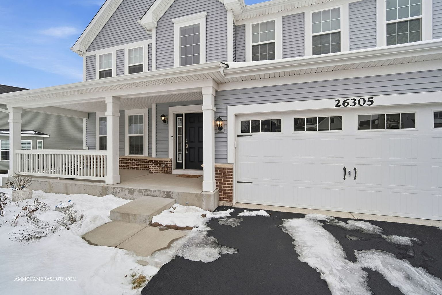 This is a front exterior view of a two-story house with light gray siding and white trim. The house features a covered front porch with white columns and a white garage door with black hardware. The address '26305' is displayed above the garage door. The driveway and front yard show patches of snow, indicating a winter setting.