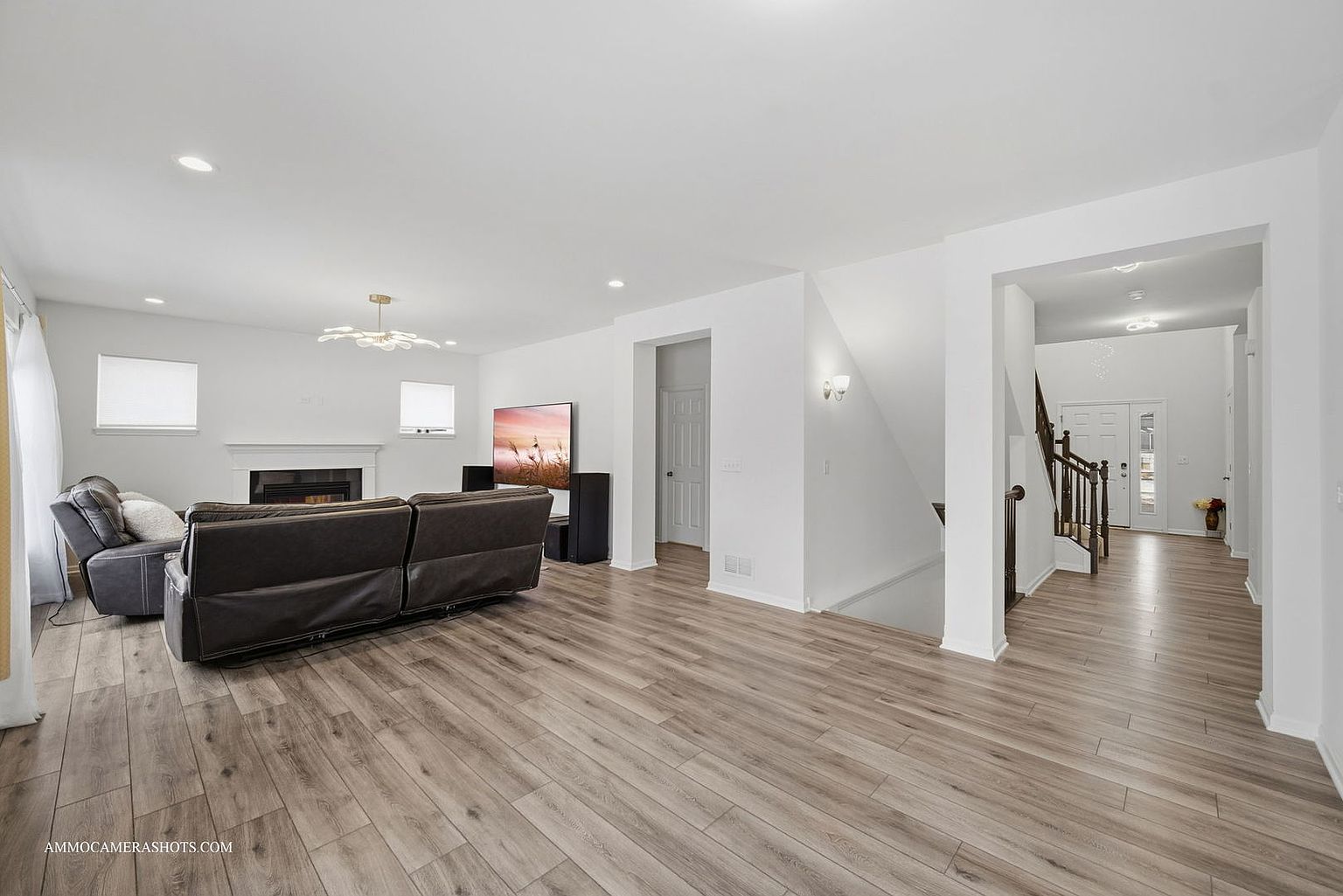 This is an interior shot of a living room featuring hardwood floors, a dark leather sofa, and a fireplace. The room is painted in neutral tones, and there is a large television on the wall. The perspective is from the entrance of the room, showcasing the open floor plan and the transition to the hallway.