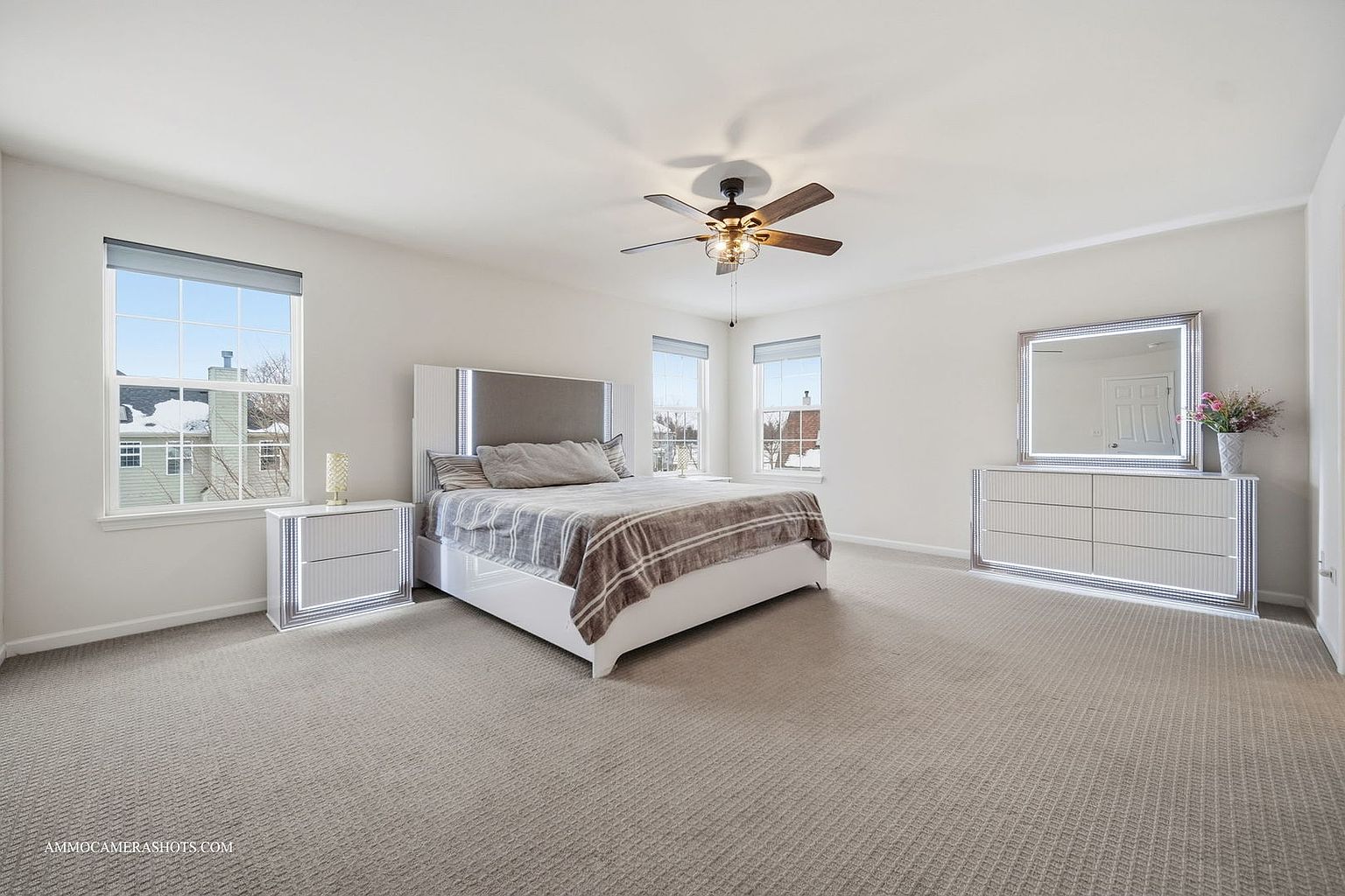 This is a bright and spacious primary bedroom featuring a large bed with a white frame and a gray headboard. The room is well-lit with natural light from multiple windows and features a ceiling fan. The furniture includes matching nightstands and a dresser with a mirror, all with modern, illuminated accents, creating a luxurious and comfortable atmosphere.