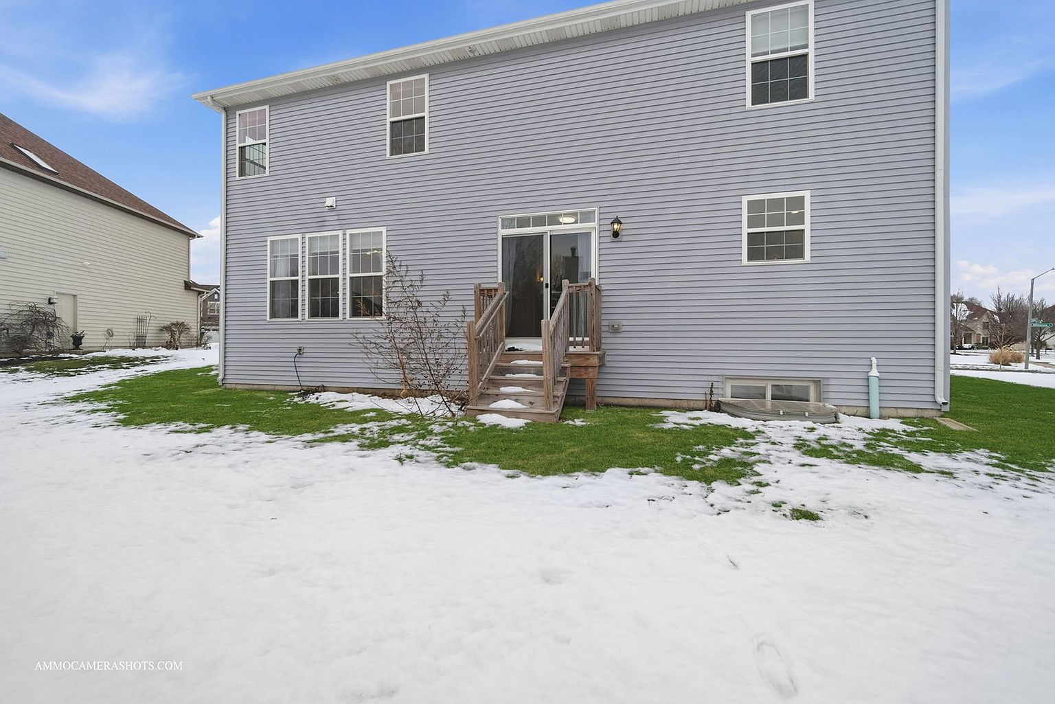 This is a rear view of a two-story house with light gray siding. A wooden deck with stairs leads to a sliding glass door. The yard is partially covered in snow, with patches of green grass visible, and a small tree is planted near the house.
