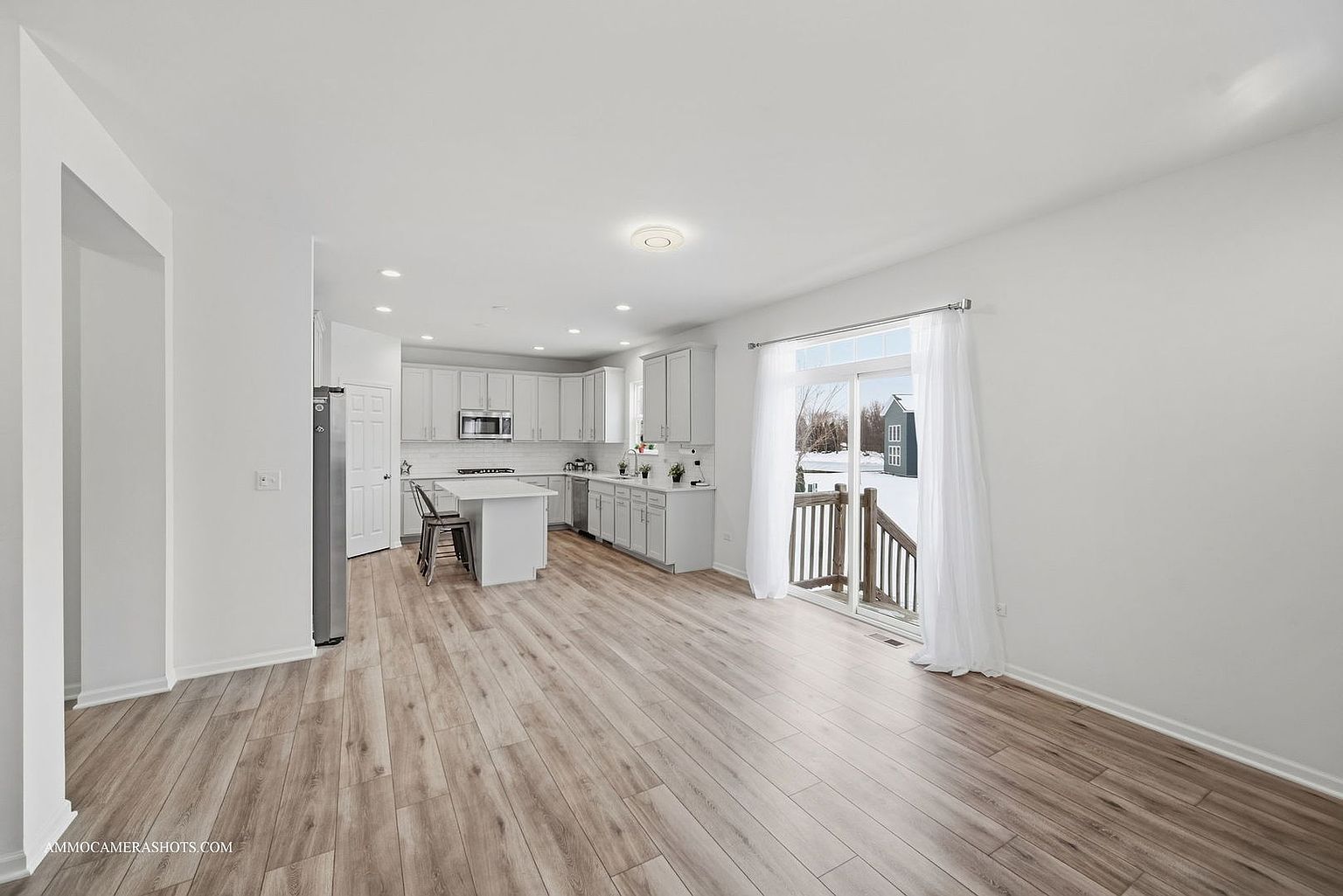 This is a bright and airy kitchen and living area featuring light wood flooring and white walls. The kitchen has white cabinets, stainless steel appliances, and a center island with seating. A sliding glass door leads to an outdoor deck, providing ample natural light and a seamless transition to outdoor living.