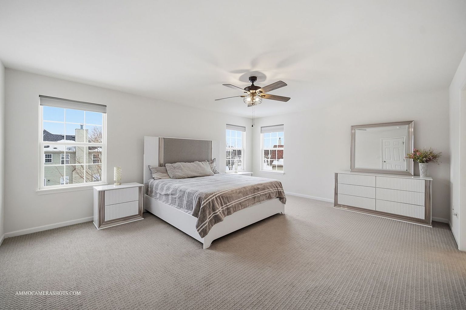 This is a bright and spacious primary bedroom featuring neutral tones and modern furnishings. The room includes a large bed with a gray and white striped blanket, a matching dresser with a mirror, and two bedside tables. Natural light floods the room through multiple windows, enhancing the airy and inviting atmosphere.