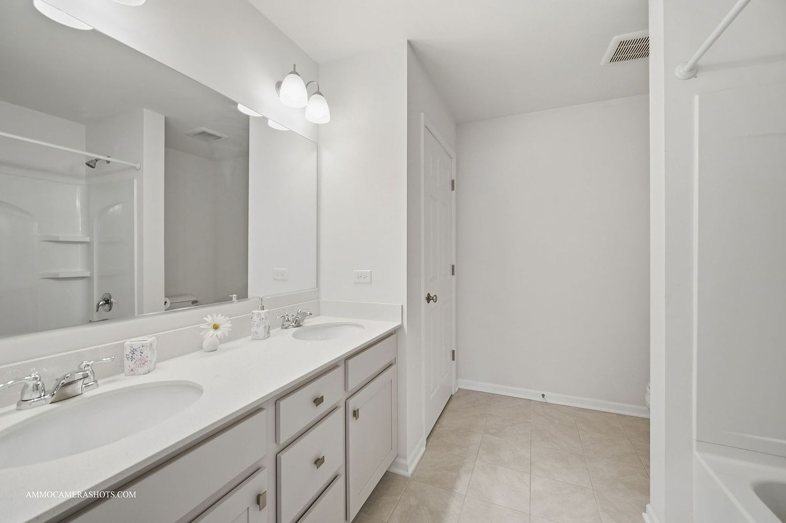 This is a well-lit bathroom featuring a double vanity with white countertops and cabinetry. A large mirror spans the length of the vanity, reflecting the shower area. The floor is tiled, and the walls are painted a clean white, creating a bright and airy atmosphere.