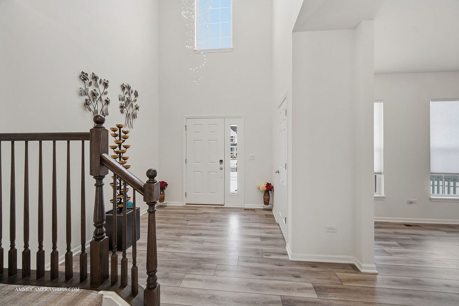 This interior shot showcases a bright and spacious entryway featuring light gray wood flooring and white walls. A dark wood staircase with decorative metal wall art adds a touch of elegance, while a high ceiling with a window allows ample natural light to flood the space. The overall impression is clean, modern, and inviting.