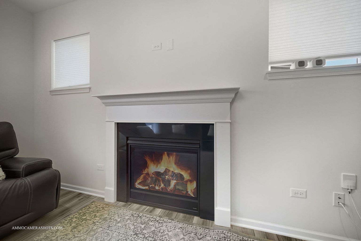 The image showcases a cozy living room interior, featuring a modern fireplace with a white mantel and a black surround. A comfortable leather recliner is partially visible on the left, and a neutral-toned rug adds warmth to the space. The room is painted in a light color, creating a bright and inviting atmosphere.