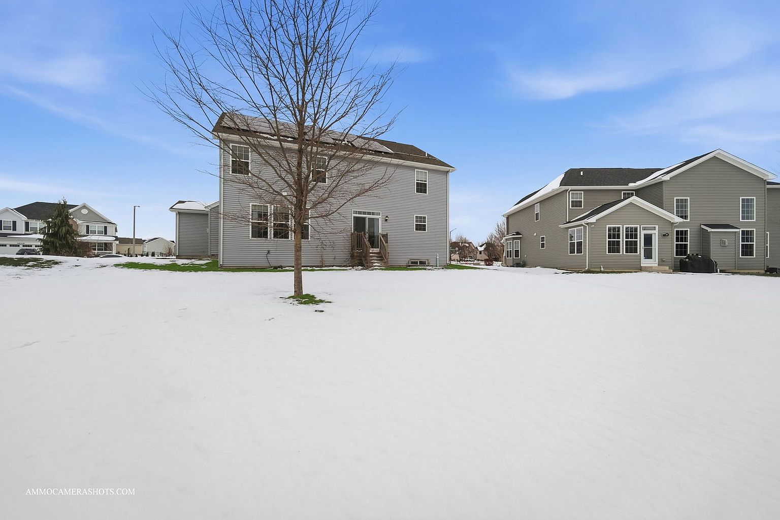 This is a rear view of a two-story house with light gray siding and a dark roof, set against a clear blue sky. The backyard is covered in snow, and a bare tree stands in the foreground. Another similar house is visible to the right, suggesting a suburban neighborhood setting.
