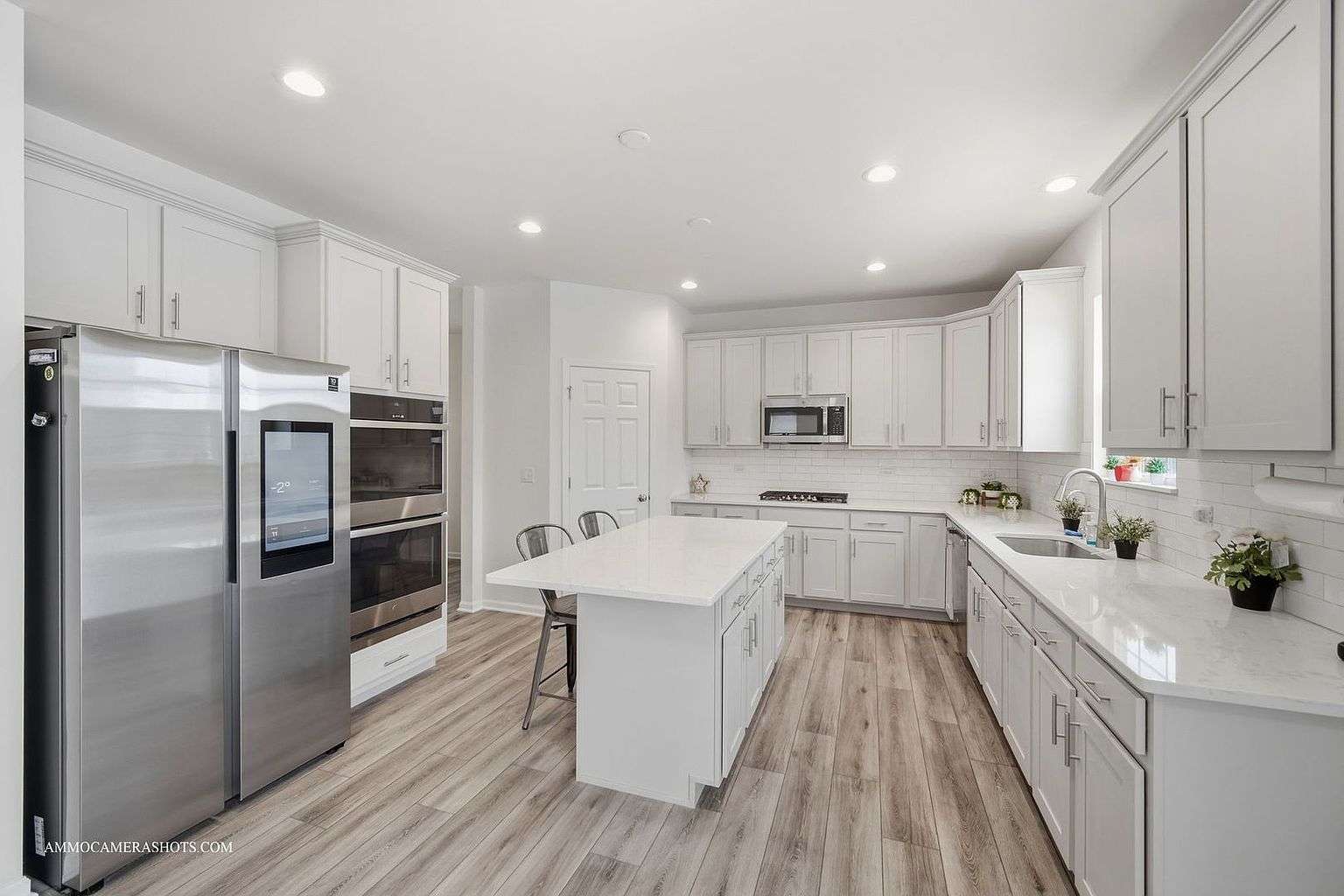 This is a bright and modern kitchen featuring white cabinets, stainless steel appliances, and light wood-look flooring. A large island with a white countertop provides ample workspace and seating. The kitchen is well-lit with recessed lighting, creating a clean and inviting atmosphere.
