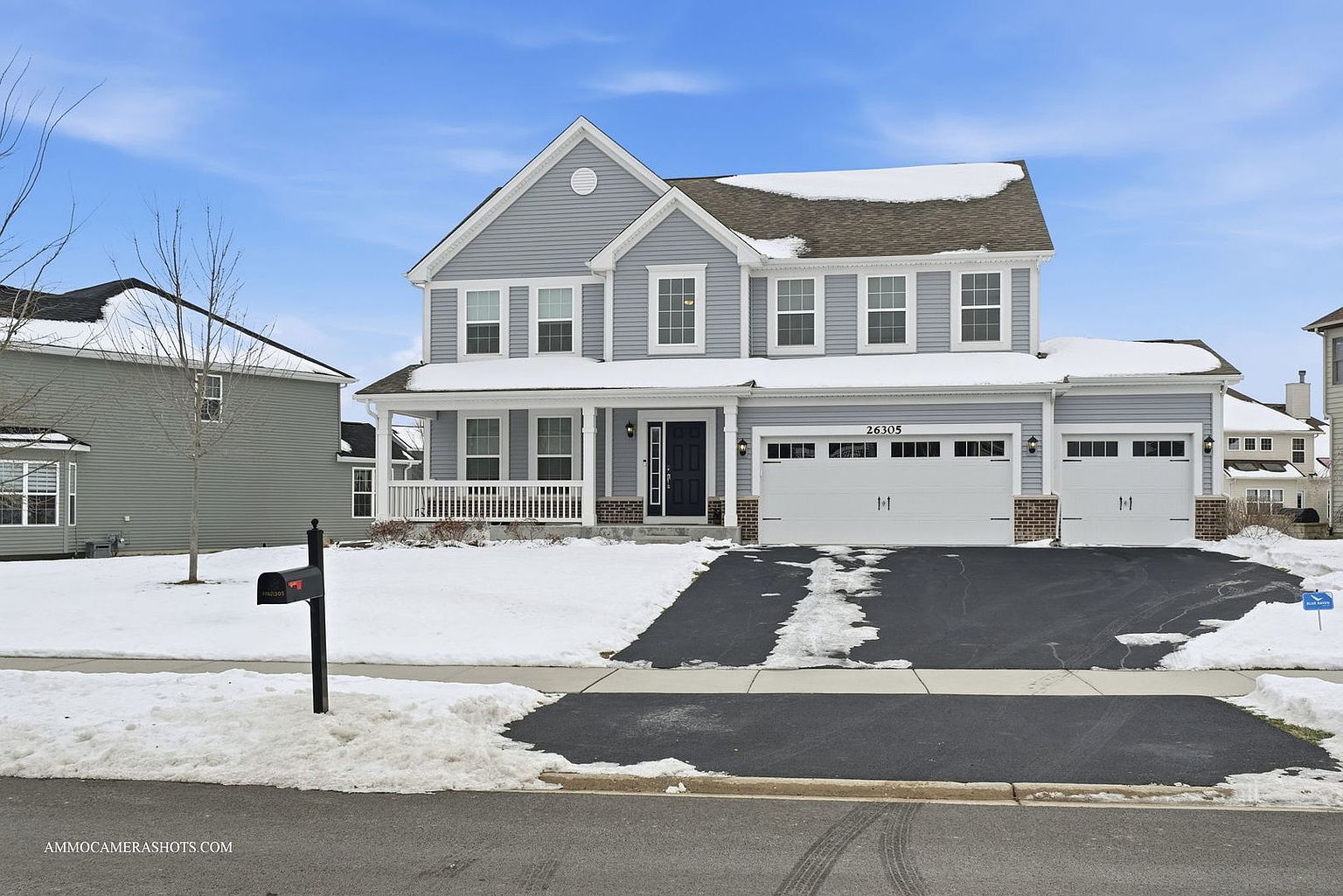 This is a front exterior view of a two-story house with a three-car garage. The house has light gray siding, white trim, and a dark front door. A snow-covered lawn and driveway add a seasonal touch, while the covered porch offers a welcoming entrance. The overall impression is a well-maintained and attractive suburban home.