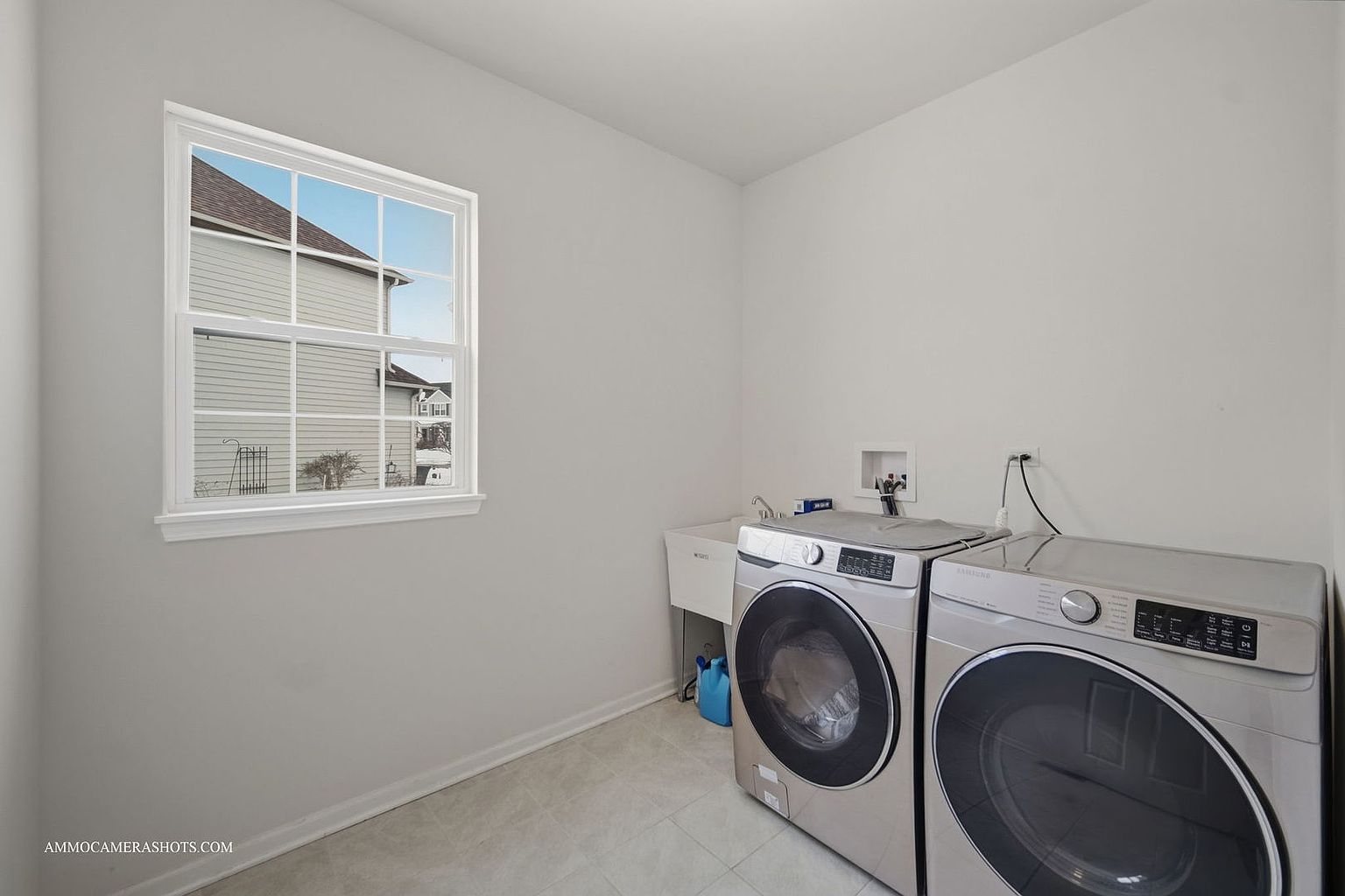 This is a well-lit laundry room featuring a front-loading washer and dryer set in a light metallic finish. A utility sink is positioned next to the washer, and a window provides natural light. The room has a clean and functional design, perfect for a modern home.