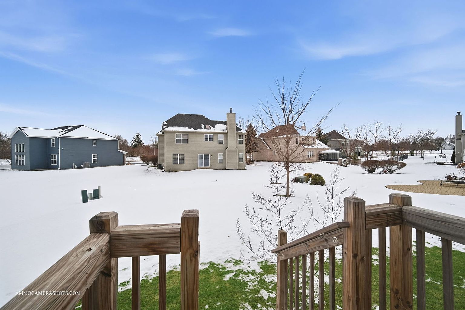 This exterior shot showcases the rear of a home with a snow-covered yard. The view is captured from a raised deck, with wooden railings framing the scene. Several houses are visible in the background, suggesting a suburban neighborhood setting.