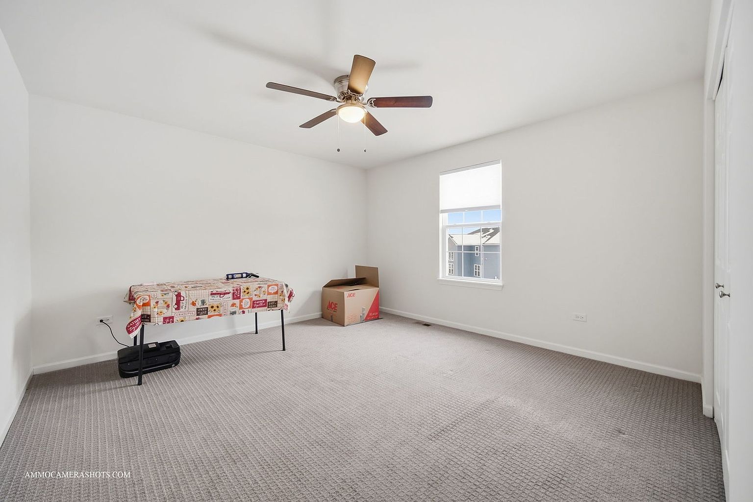 This is a bright and spacious bedroom featuring neutral carpet and white walls, creating a clean and inviting atmosphere. A ceiling fan provides air circulation, while a window allows natural light to fill the room. The room is sparsely furnished with a table covered with a decorative cloth and a cardboard box, suggesting it's staged or awaiting personalization.