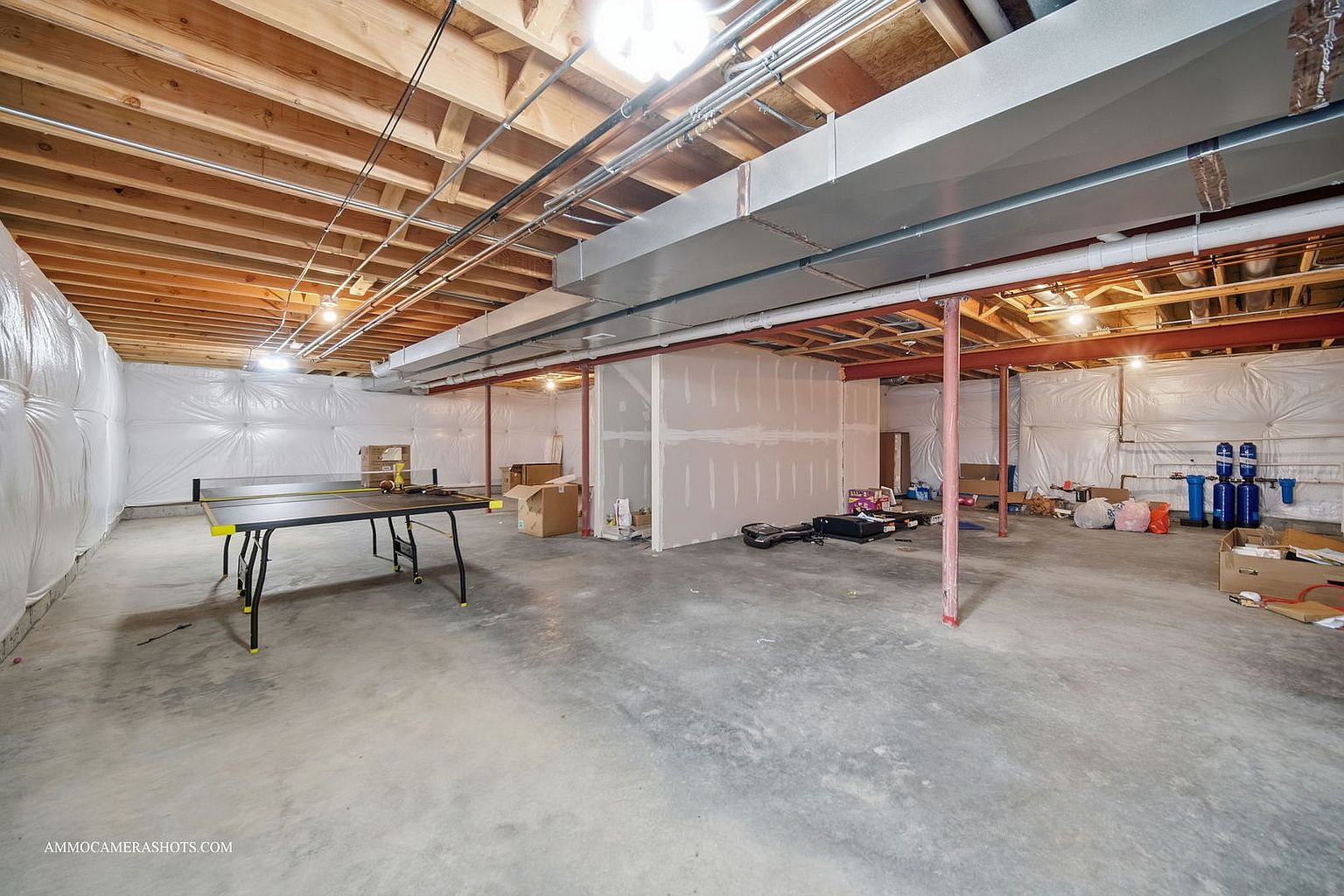This is a spacious unfinished basement with exposed wooden ceiling beams and ductwork. The concrete floor is expansive, and the walls are partially covered with white plastic sheeting and drywall. A ping pong table sits on the left side, and various boxes and items are scattered throughout the space, suggesting potential for storage or customization.