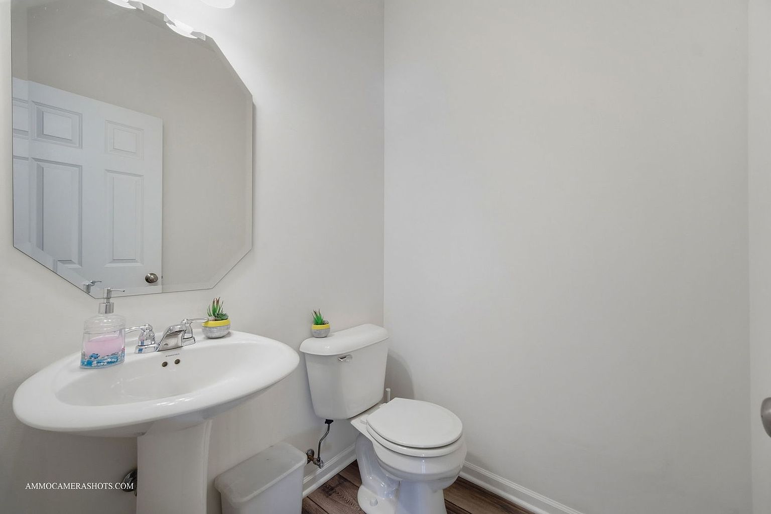 This is a well-lit guest bathroom featuring a pedestal sink with a unique mirror above it, a standard toilet, and neutral-colored walls. The flooring appears to be wood-look laminate, adding a touch of warmth to the space. The room is clean and functional, ideal for a half-bath or powder room.