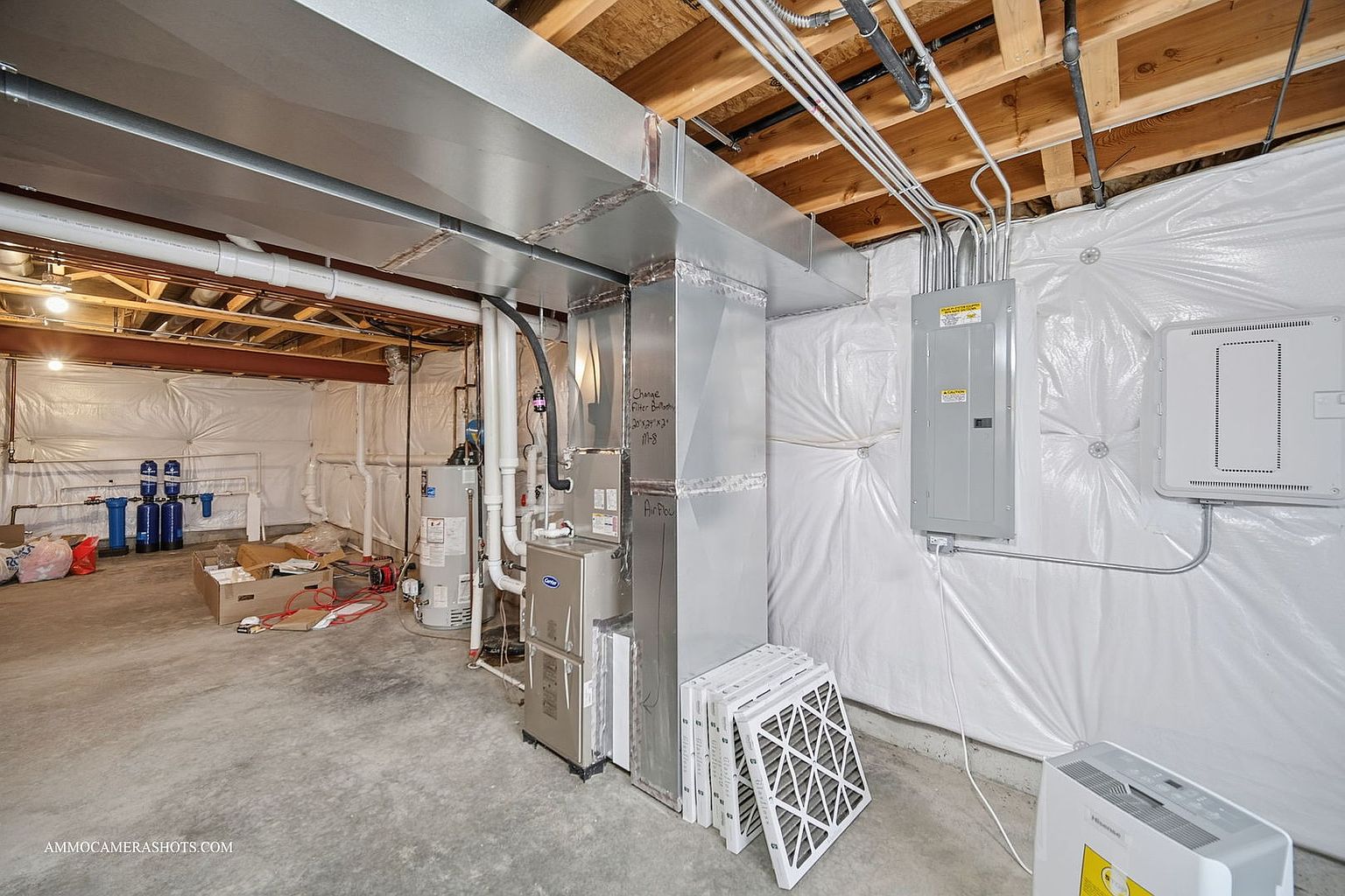 This is a basement interior featuring essential utilities and a clean, unfinished aesthetic. The space includes a furnace, water heater, electrical panel, and ductwork, all neatly arranged against a backdrop of white plastic sheeting on the walls. The concrete floor is bare, suggesting a functional, utilitarian area ready for customization or storage.