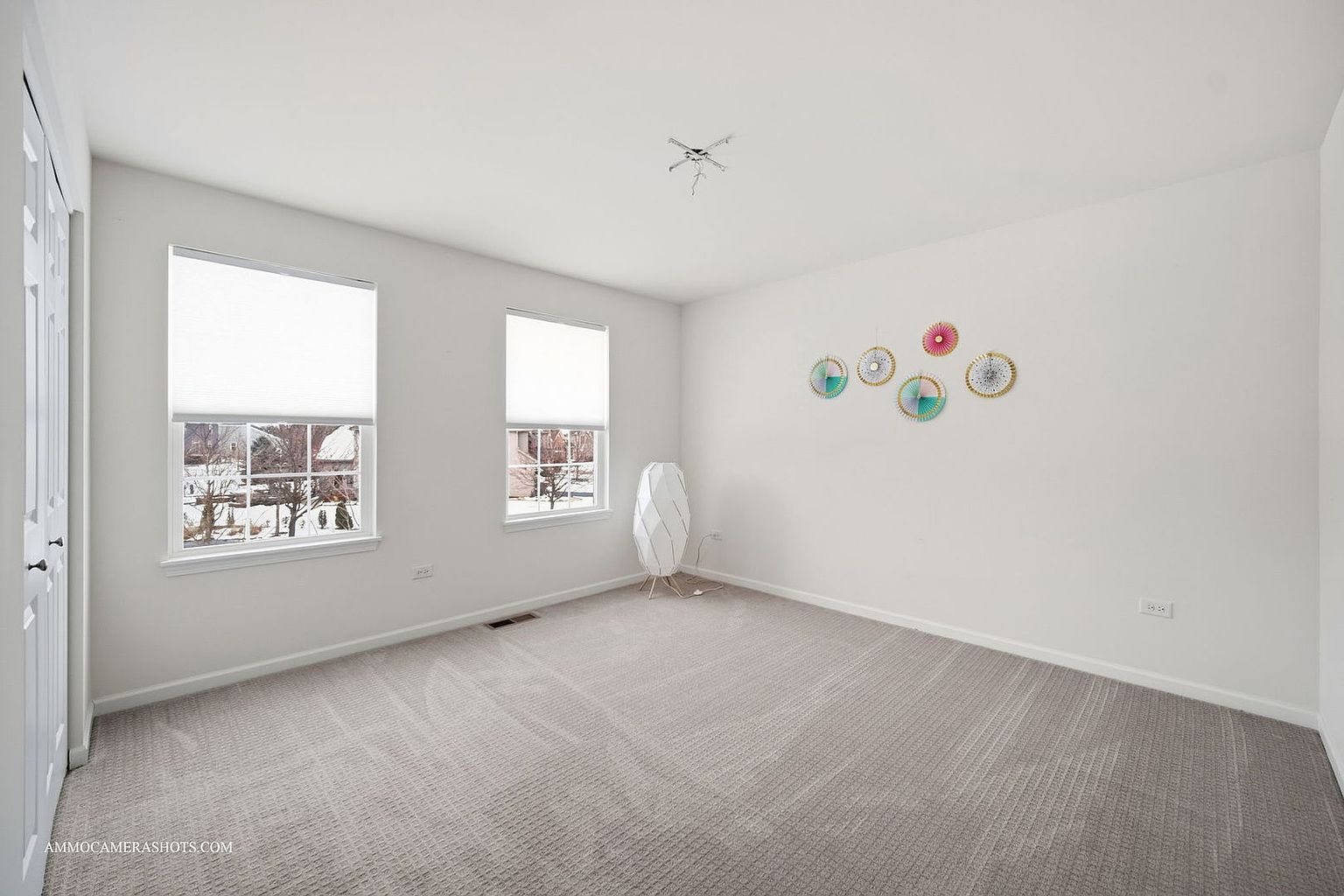 This is a bright and airy bedroom featuring neutral gray carpet and white walls. Two windows with white blinds provide natural light, and decorative wall art adds a touch of color. A modern floor lamp stands near one of the windows, and the room appears clean and well-maintained, offering a blank canvas for potential buyers.
