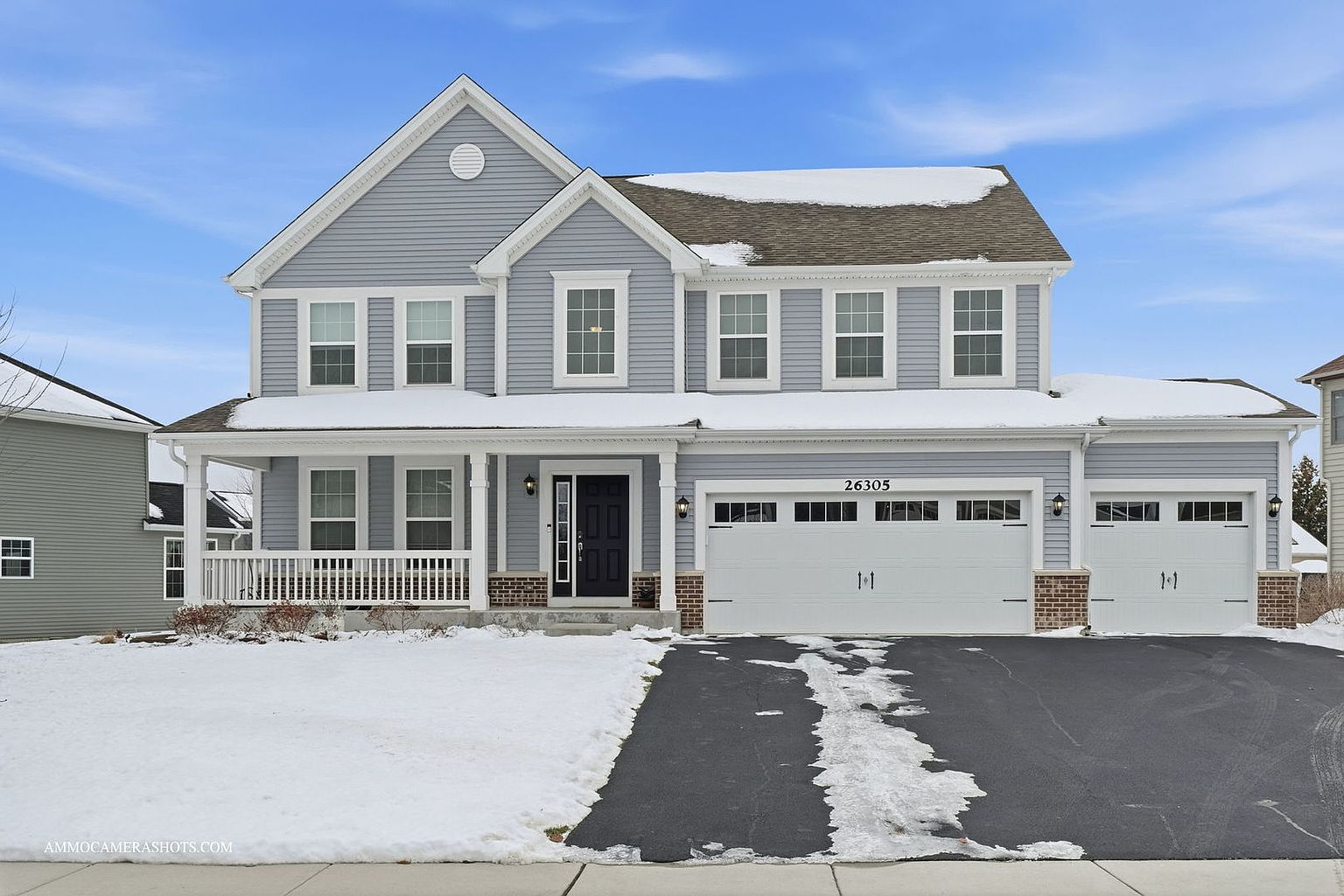 This is a front exterior view of a two-story house with light blue siding and white trim. The house features a covered front porch, a two-car garage, and a well-maintained lawn partially covered in snow. The driveway is paved and leads up to the garage, and the house number '26305' is visible above the garage door.
