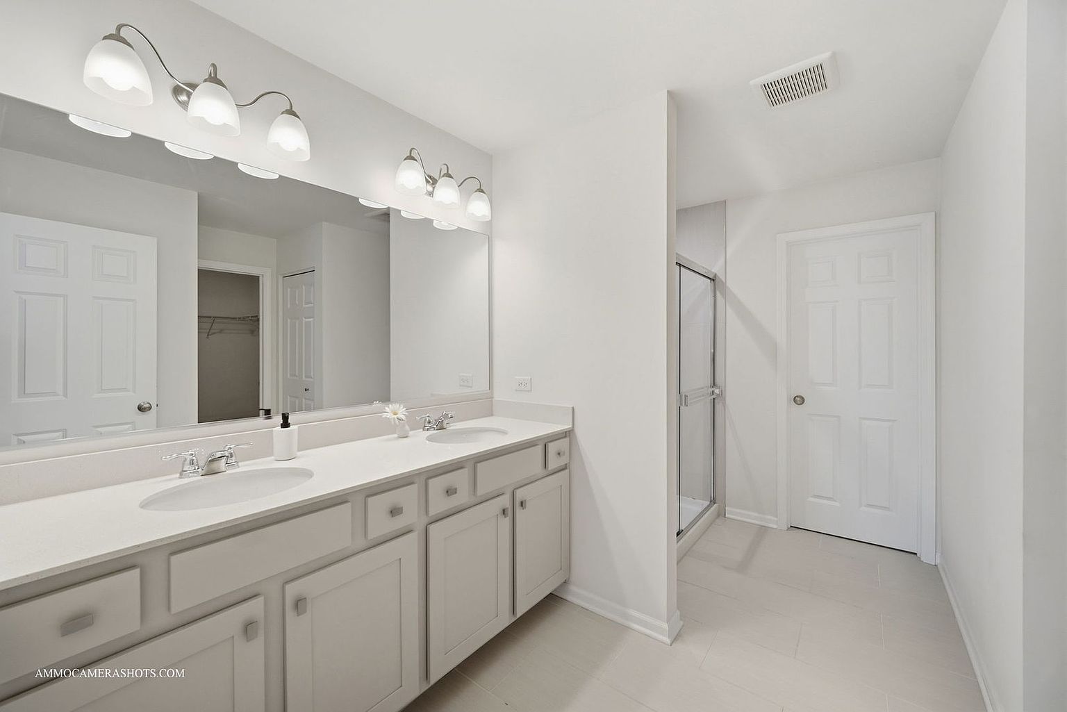This is a bright and clean primary bathroom featuring a double vanity with white countertops and light gray cabinetry. A large mirror spans the length of the vanity, illuminated by modern light fixtures. The bathroom also includes a glass-enclosed shower and a white door, contributing to the room's fresh and airy feel.