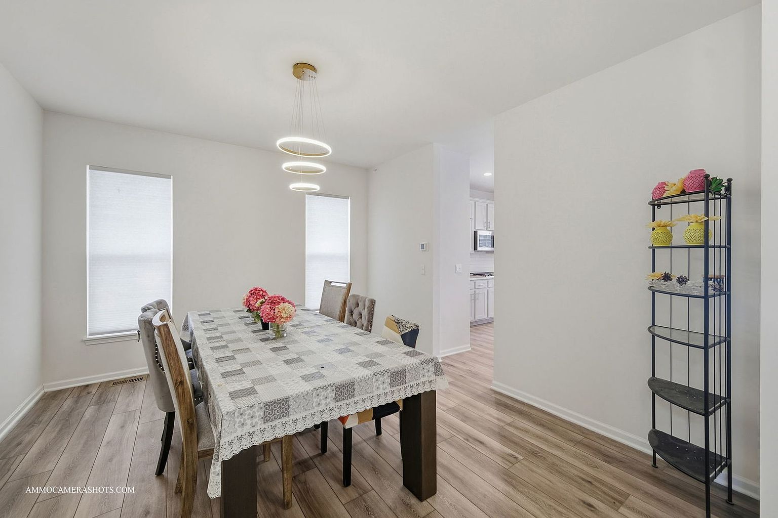 This is an interior shot of a dining room featuring a long dining table covered with a patterned tablecloth and surrounded by chairs. A modern, circular chandelier hangs above the table, and natural light streams in through two windows with blinds. A tall, decorative shelving unit stands against the wall, adding a touch of elegance to the space.