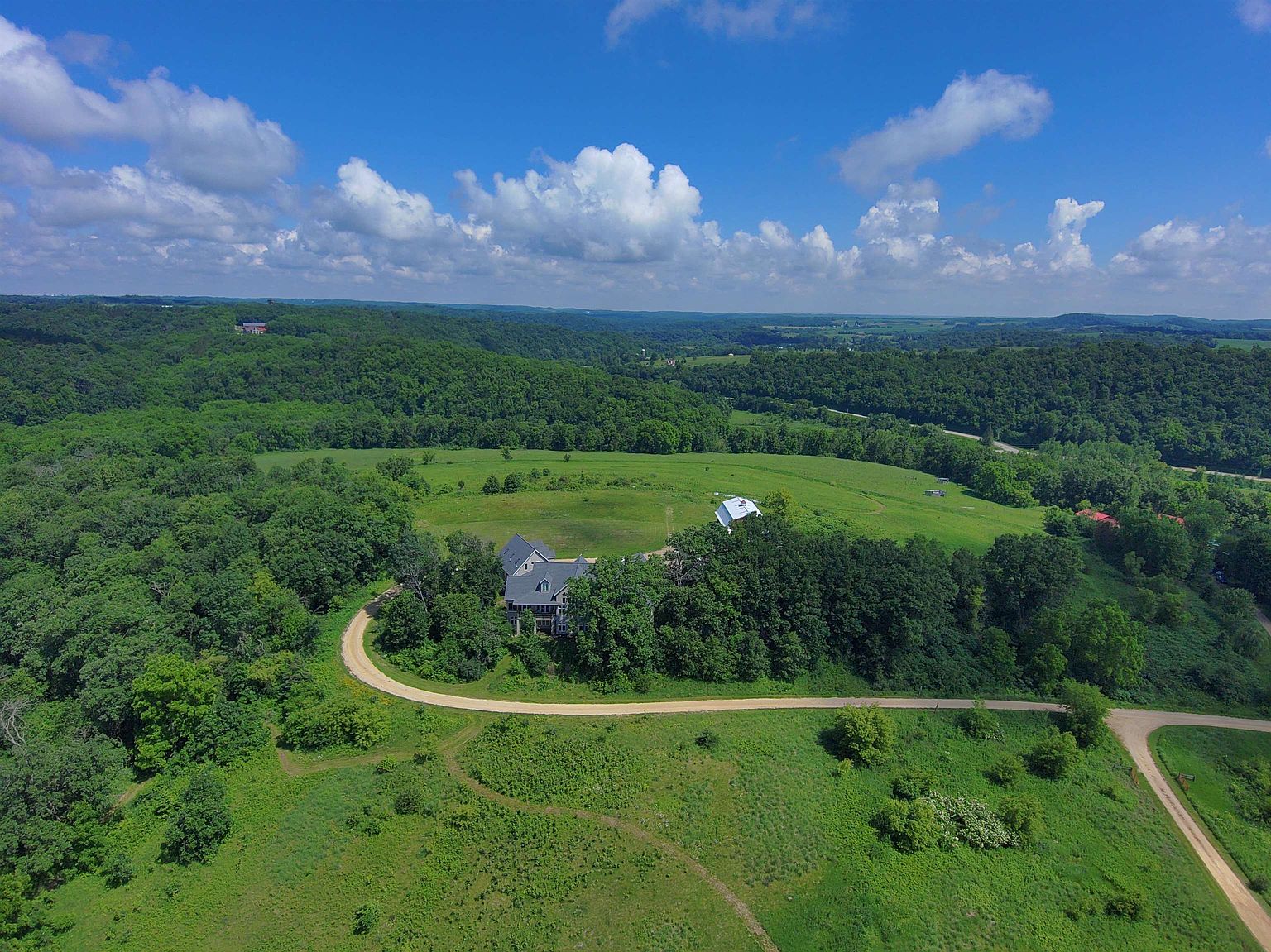 This high-angle aerial shot captures a secluded residential property nestled within a lush, rolling landscape of dense forests and open green fields. A winding gravel driveway leads to a multi-story home with a dark-toned roof, surrounded by mature trees that provide privacy and shade. The expansive, picturesque setting emphasizes the property's rural charm and connection to the surrounding natural environment.