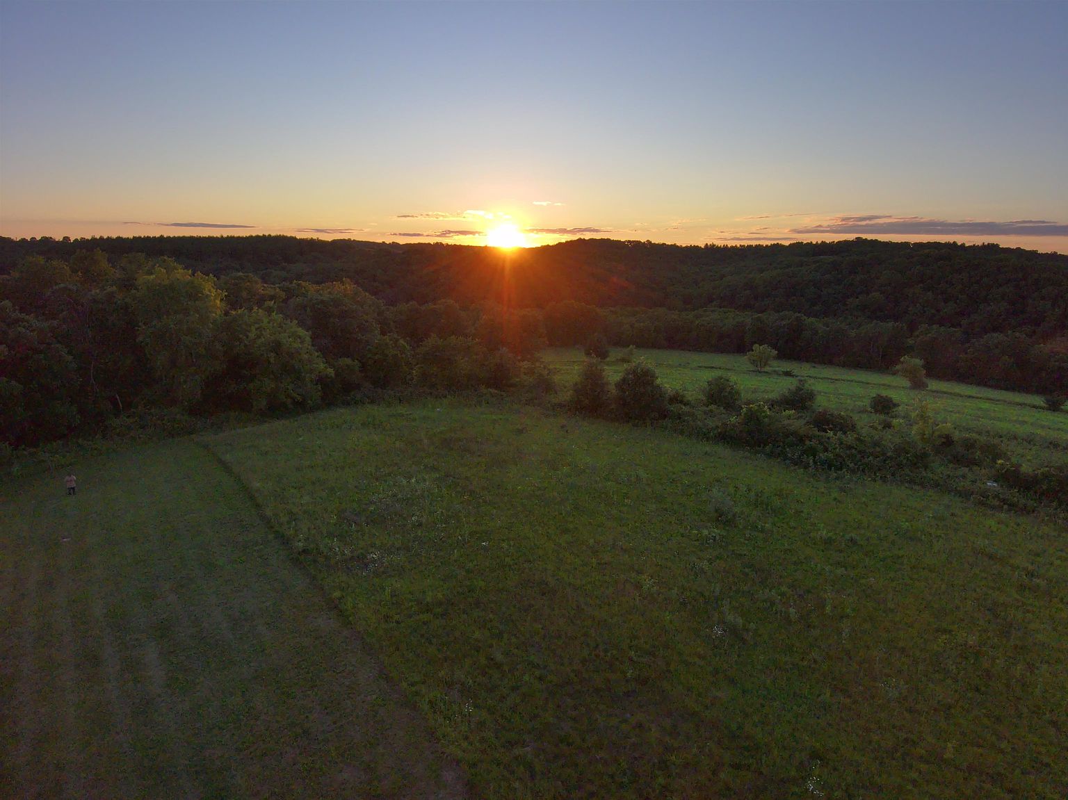 This aerial perspective captures a vast, rolling landscape at sunset, showcasing a large, open grassy field that transitions into a dense, forested hillside. The sun sits low on the horizon, casting a warm, golden glow across the terrain and creating a dramatic lens flare effect. The expansive view highlights the natural beauty and privacy of the property, emphasizing its serene and rural setting.