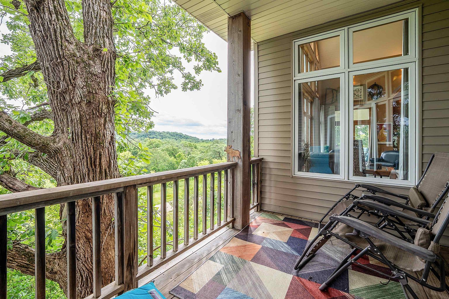 This elevated wooden deck offers a serene outdoor living space with a scenic view of lush, rolling hills and dense forest. The area features a geometric patterned rug, two reclining lounge chairs, and a wooden railing that integrates with the surrounding mature trees. Large windows on the adjacent wall provide a seamless connection to the interior living space, creating a peaceful and inviting atmosphere.
