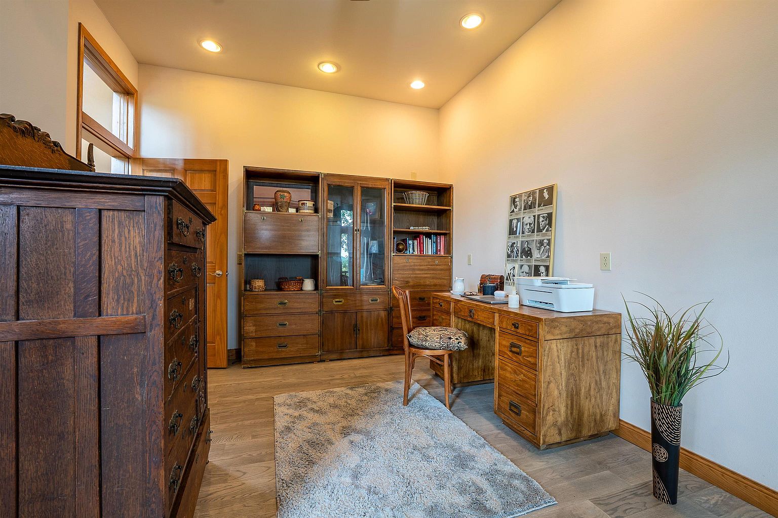 This home office features a warm, traditional aesthetic with rich wood furniture, including a large desk and a matching multi-section wall unit. A patterned area rug sits on the light-toned flooring, while a framed collection of portraits adds a classic touch to the wall. The room is illuminated by recessed ceiling lights, creating a professional and inviting workspace.