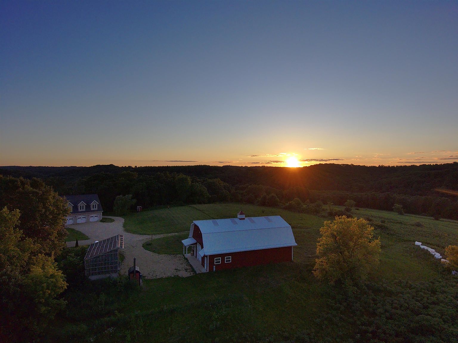 This aerial perspective captures a serene rural property at sunset, featuring a classic red barn with a white roof as the central focus. To the left, a multi-car garage building sits adjacent to a gravel driveway, surrounded by expansive green fields and rolling hills. The warm, golden light of the setting sun creates a peaceful and picturesque atmosphere, highlighting the vast landscape and the property's quiet, countryside setting.