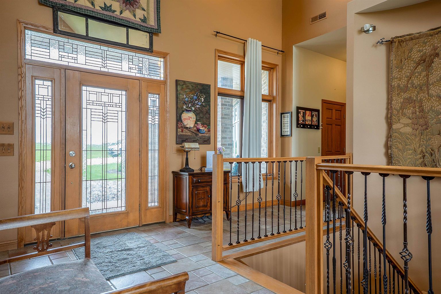This inviting entryway features a grand wooden front door with intricate glass panels, allowing natural light to fill the space. A wooden console table sits against the wall beneath a framed floral artwork, while a staircase with decorative metal balusters leads to a lower level. The warm, neutral wall tones and tiled flooring create a welcoming and traditional atmosphere.