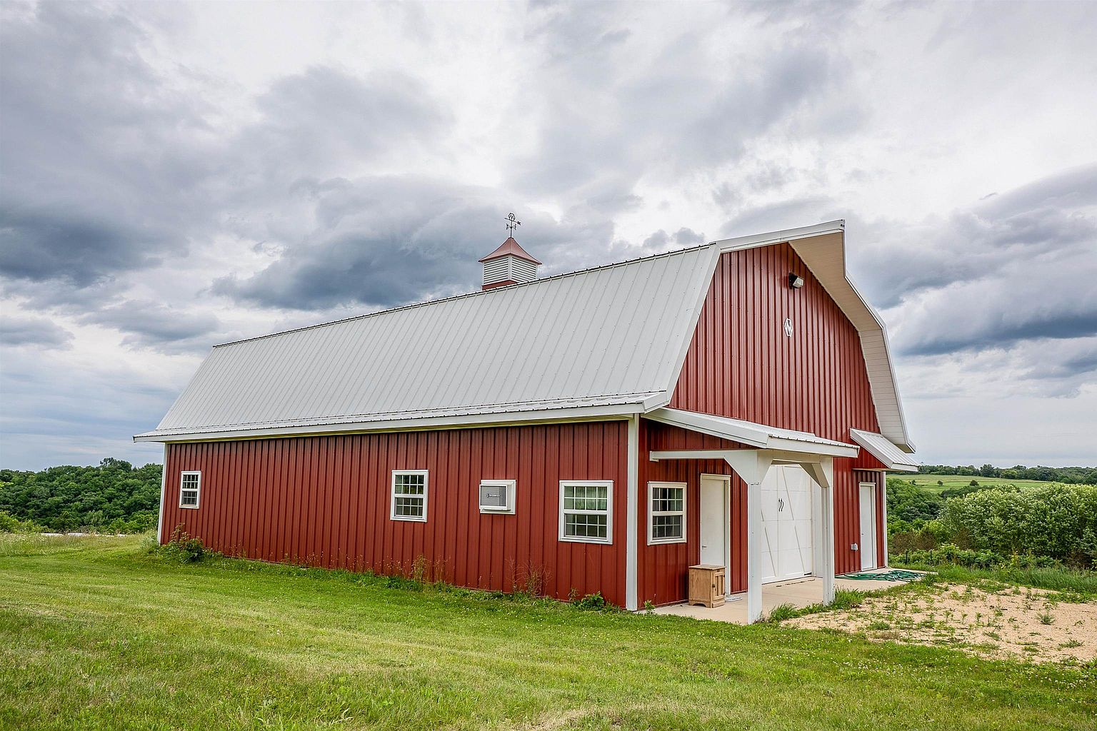 This image captures a side view of a large, red metal-sided barn with a white metal roof, set against a cloudy, dramatic sky. The structure features a cupola on the roof, multiple grid-patterned windows along the side, and a covered entryway with a white support post. The barn is situated on a grassy, slightly sloped lot with a gravel area near the entrance, suggesting a rural or agricultural property setting.