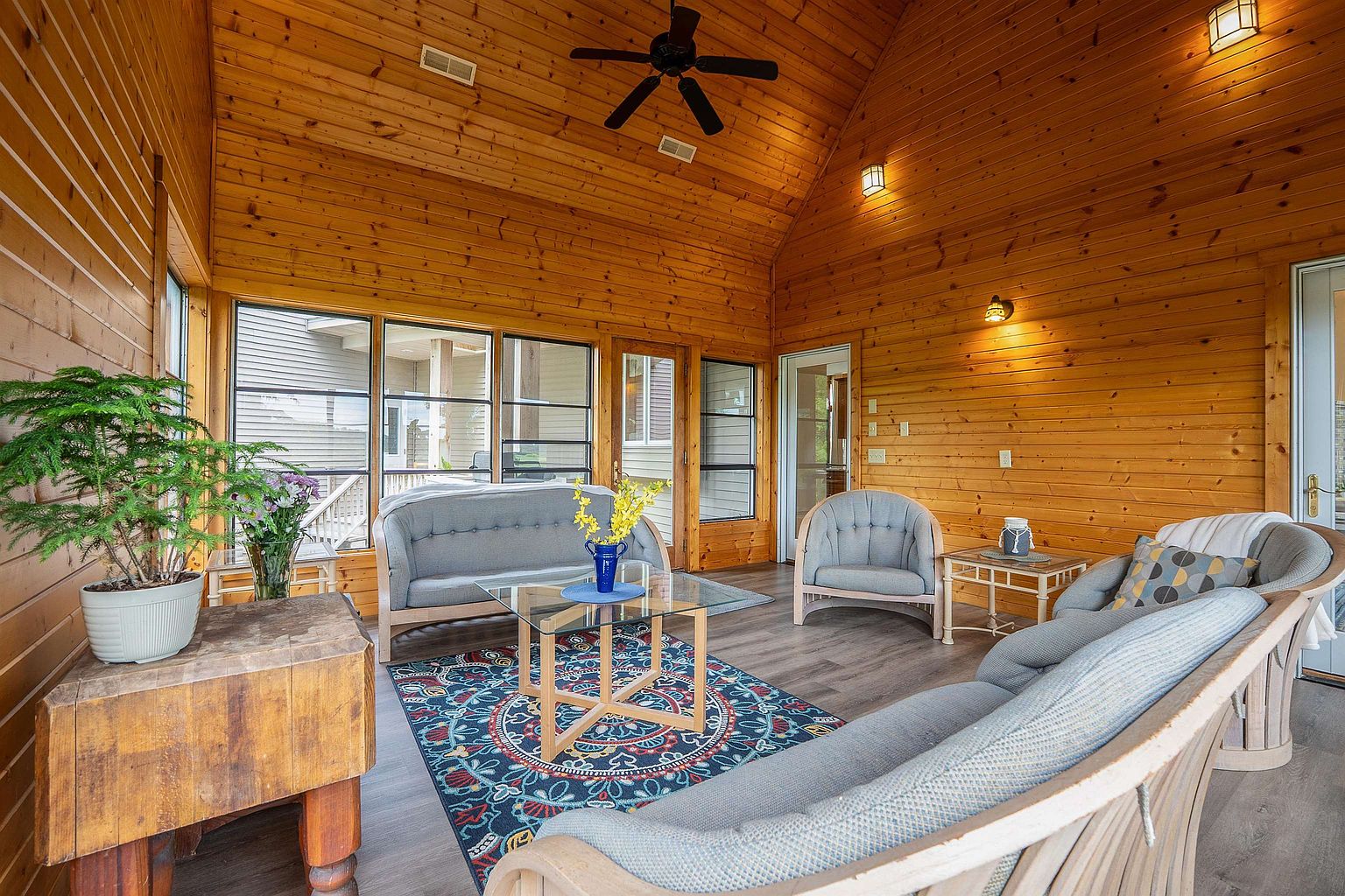 This bright and airy sunroom features warm wood-paneled walls and a vaulted ceiling with a central fan, creating a cozy, rustic atmosphere. The space is furnished with a light gray sofa, matching armchair, and a glass-topped coffee table centered on a vibrant, patterned area rug. Large windows provide ample natural light and views of the exterior, while a sturdy wooden block table in the foreground adds a unique, textured element to the room.