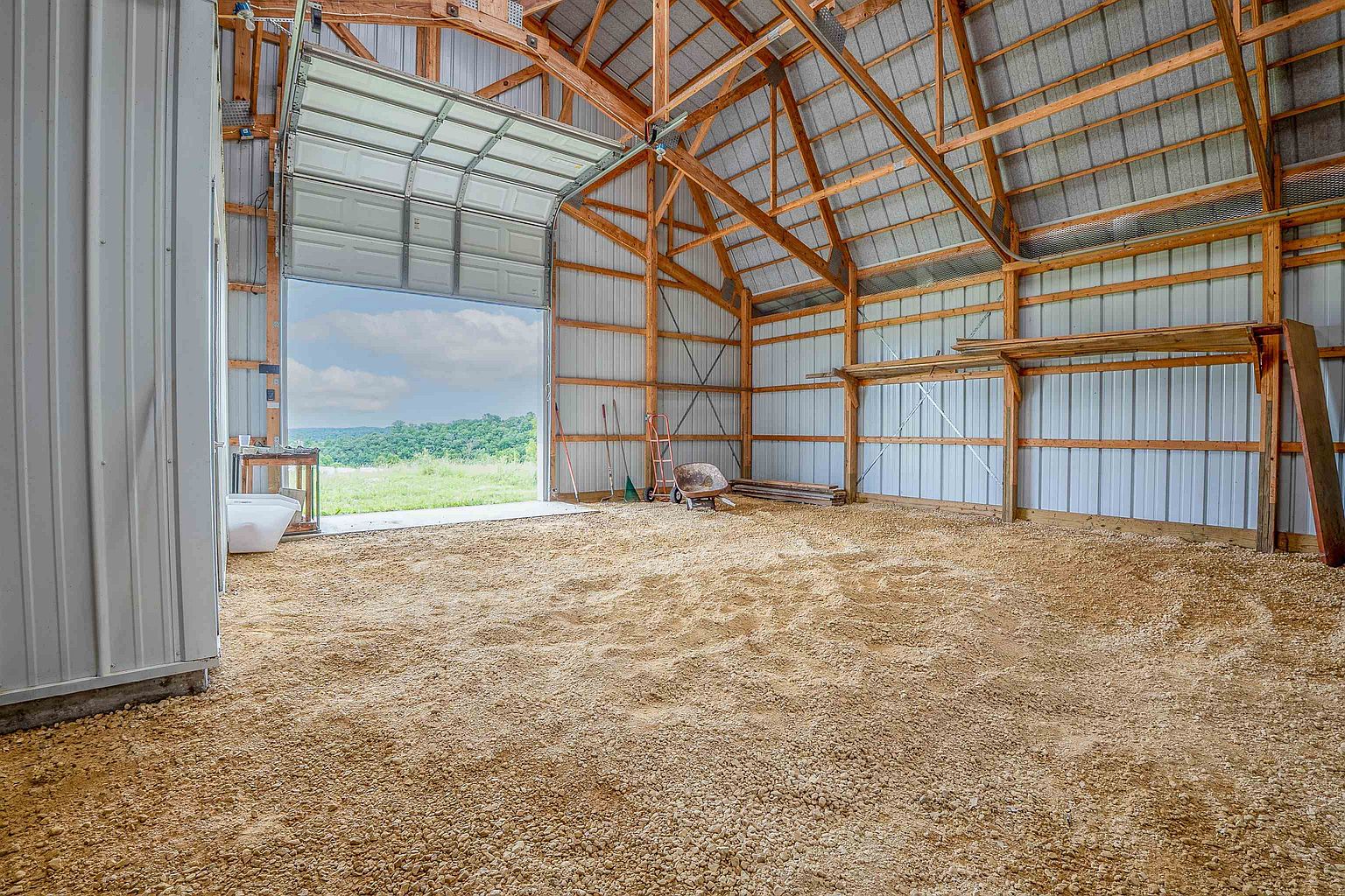 This image shows the interior of a large, open-concept barn or workshop with a gravel floor and exposed wooden framing. A large overhead garage door is partially open, revealing a scenic view of a grassy field and distant hills. The structure features metal wall panels and a high, vaulted ceiling, providing a spacious and versatile area for storage or projects.