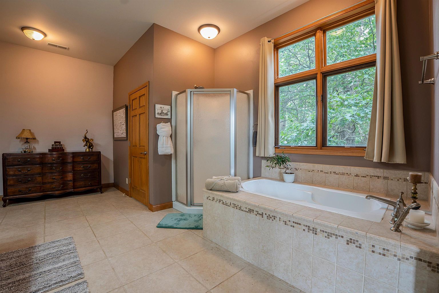 This spacious primary bathroom features a large soaking tub with a tiled surround positioned beneath a window that looks out onto lush greenery. A corner glass-enclosed shower stands adjacent to a wooden dresser that serves as a unique storage piece, while neutral-toned floor tiles ground the space. The room is illuminated by two ceiling-mounted light fixtures, creating a warm and inviting atmosphere.