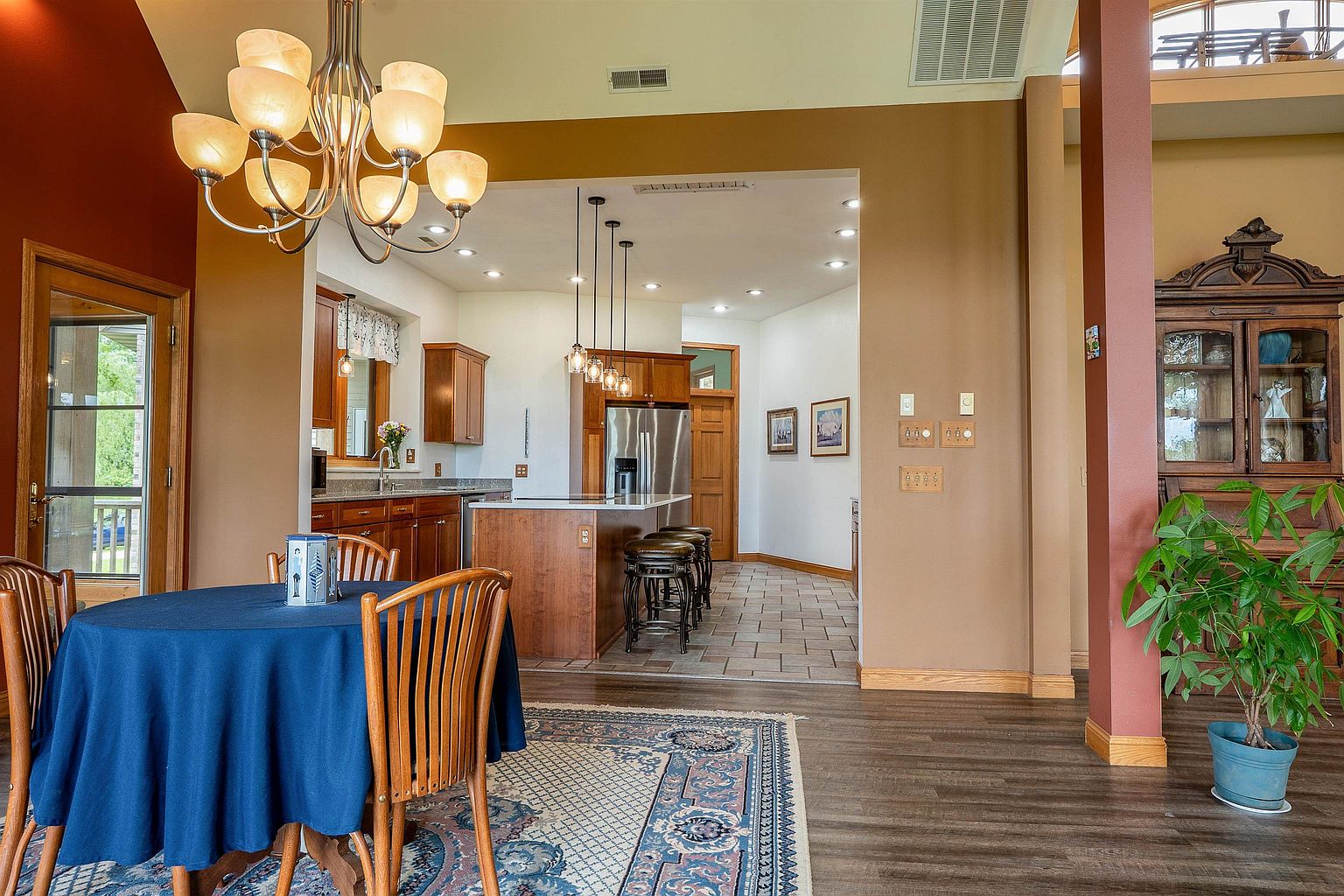 This view captures a dining area in the foreground that transitions into an open-concept kitchen. The dining space features a round table with a blue cloth and a decorative chandelier, while the kitchen beyond showcases dark wood cabinetry, a central island with bar seating, and stainless steel appliances. The home has a warm, traditional aesthetic with a mix of wood-look flooring and tile, complemented by a large ornate hutch on the right.