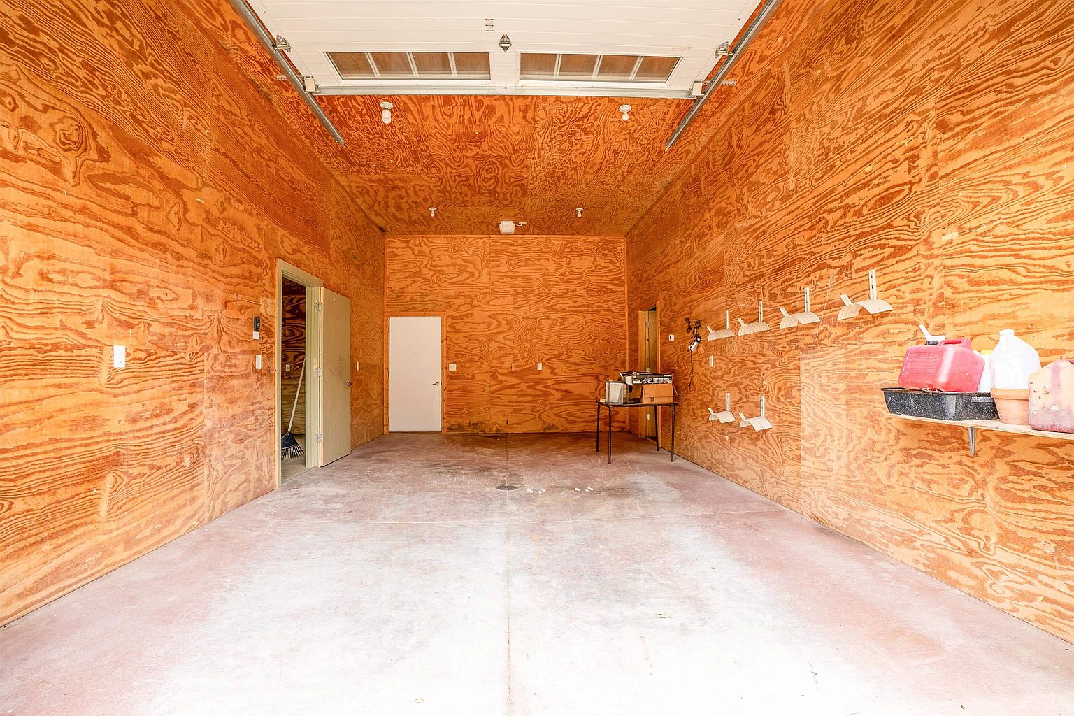This interior view of a spacious garage features unfinished plywood walls and a concrete floor, providing a rustic and functional aesthetic. The space includes a high ceiling with exposed lighting fixtures, a side door, and a wall-mounted storage rack for tools. The perspective looks straight through the garage toward the back wall, highlighting the depth and utility of the area.