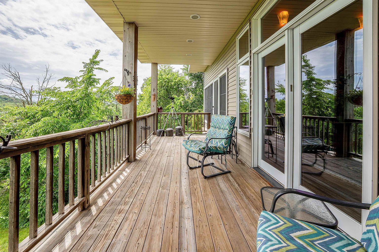 This spacious wooden deck offers a serene outdoor living space with a panoramic view of the surrounding lush green trees. The area is furnished with comfortable seating, including a patterned chair, and features a hanging planter, creating a relaxing atmosphere. Large glass doors provide seamless access from the interior, making this an ideal spot for enjoying nature and fresh air.