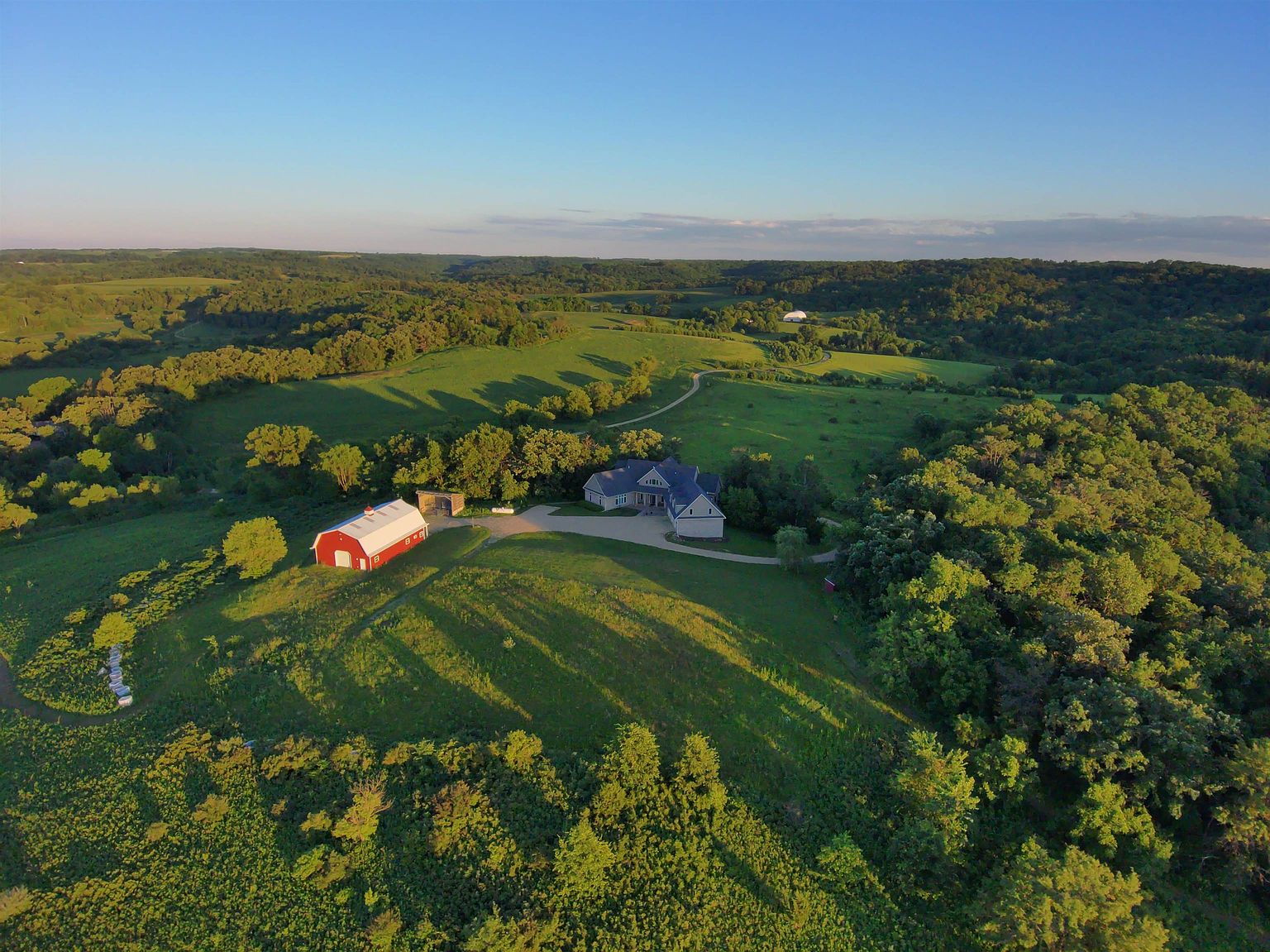 This high-angle aerial view captures a sprawling rural estate nestled within a lush, rolling landscape of green hills and dense forest. The property features a large, multi-gabled residence with a paved driveway and a distinct red barn, all bathed in the warm, golden light of late afternoon. The expansive grounds emphasize privacy and a serene, natural setting, showcasing the home's integration into the surrounding countryside.