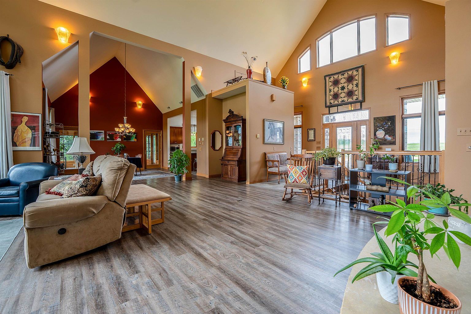 This spacious, open-concept living area features vaulted ceilings and warm, neutral-toned walls, creating an inviting and airy atmosphere. The room is anchored by wood-look flooring and includes a comfortable seating area with a plush sofa, a rocking chair, and a decorative hutch. Large windows allow natural light to fill the space, highlighting the architectural depth and the transition into the adjacent dining area.