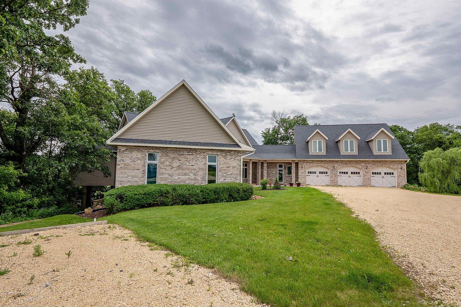 This front-facing view captures a sprawling, multi-gabled residence featuring a blend of light-colored brick and horizontal siding. The home includes a three-car garage with arched carriage-style doors and a gravel driveway that leads to a landscaped front lawn. The property is surrounded by mature trees, creating a private and serene atmosphere.