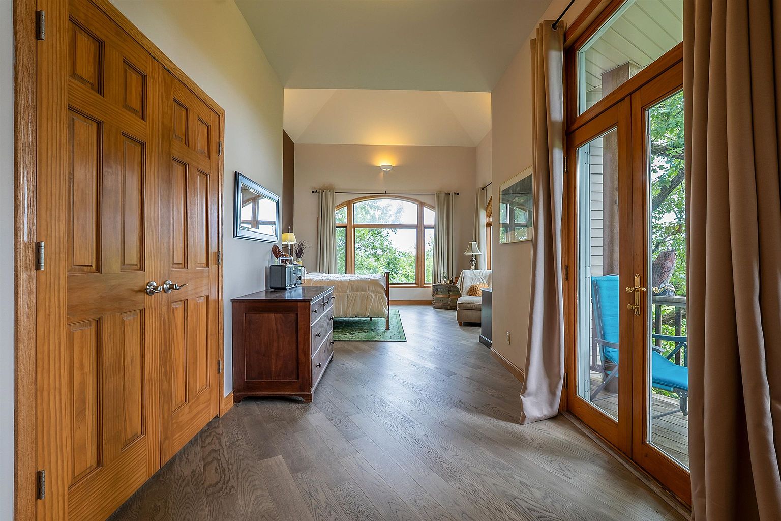 This primary bedroom suite features a spacious layout with light wood flooring arranged in a herringbone pattern. The room transitions from a hallway area with a wooden dresser into a sleeping space anchored by a large, arched window that provides ample natural light and views of the surrounding trees. To the right, glass-paned double doors open onto a private deck, creating a seamless connection between the indoor living space and the outdoors.