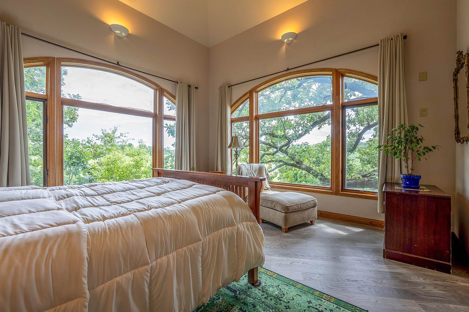 This bright and airy bedroom features a wooden bed frame centered in the room, flanked by large, arched windows that offer expansive views of the surrounding trees. A cozy armchair with an ottoman sits near the window, creating a peaceful reading nook, while a wooden dresser with a potted plant adds warmth to the space. The room is finished with light-toned flooring and a patterned area rug, providing a serene and inviting atmosphere.