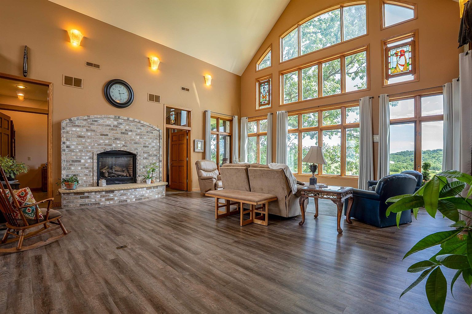 This spacious living room features a vaulted ceiling and a prominent brick fireplace with a stone hearth as its central focal point. A wall of large windows provides abundant natural light and expansive views of the surrounding landscape, complemented by stained glass accents. The room is furnished with a comfortable seating area, a wooden rocking chair, and wood-look flooring, creating a warm and inviting atmosphere.