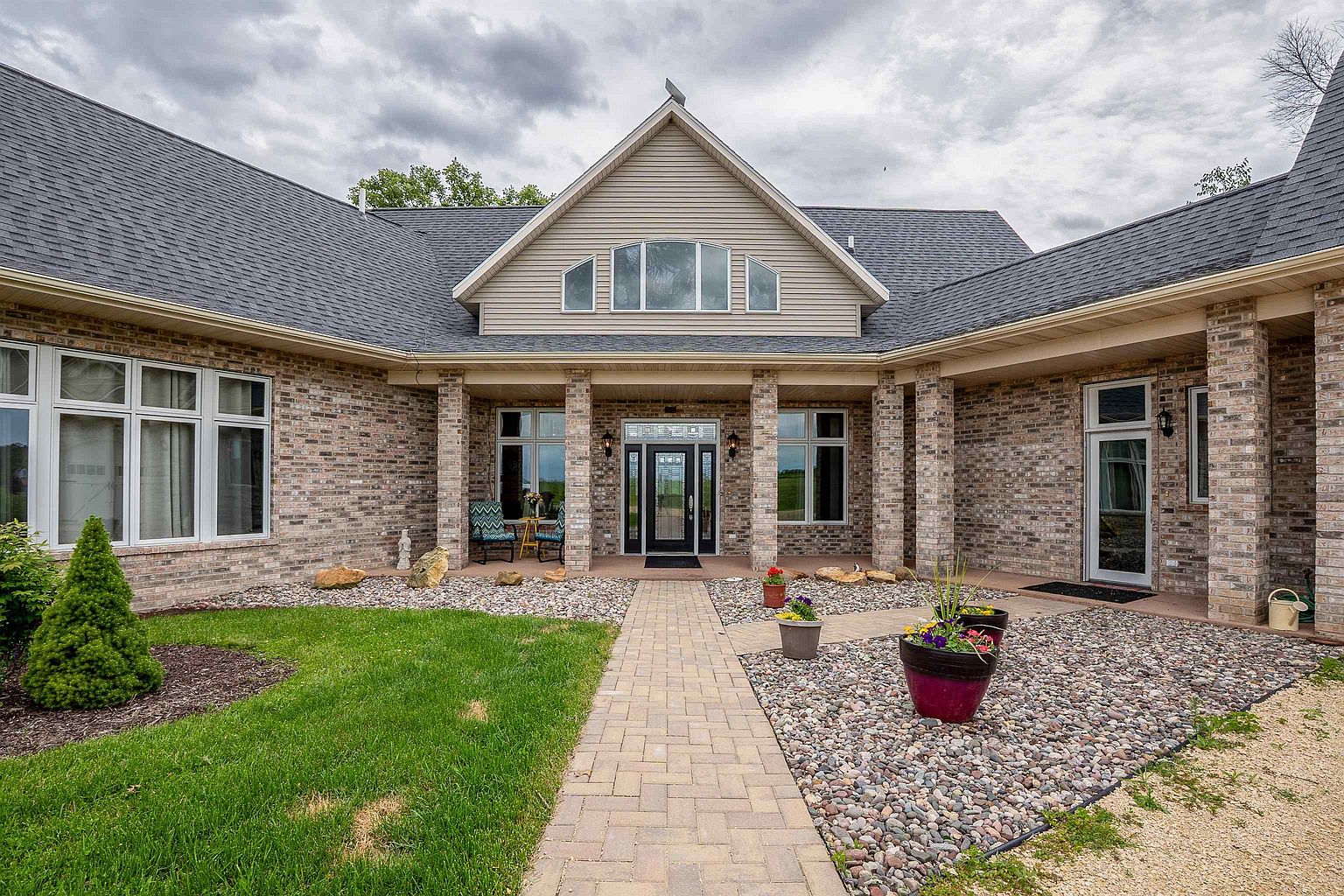 This expansive single-story residence features a classic brick facade with a multi-gabled roofline and a large gravel driveway. The home includes a three-car garage on the right and a covered entryway centered between symmetrical wings, all set against a cloudy sky. The wide-angle perspective captures the full scale of the property and its rural, open setting.
