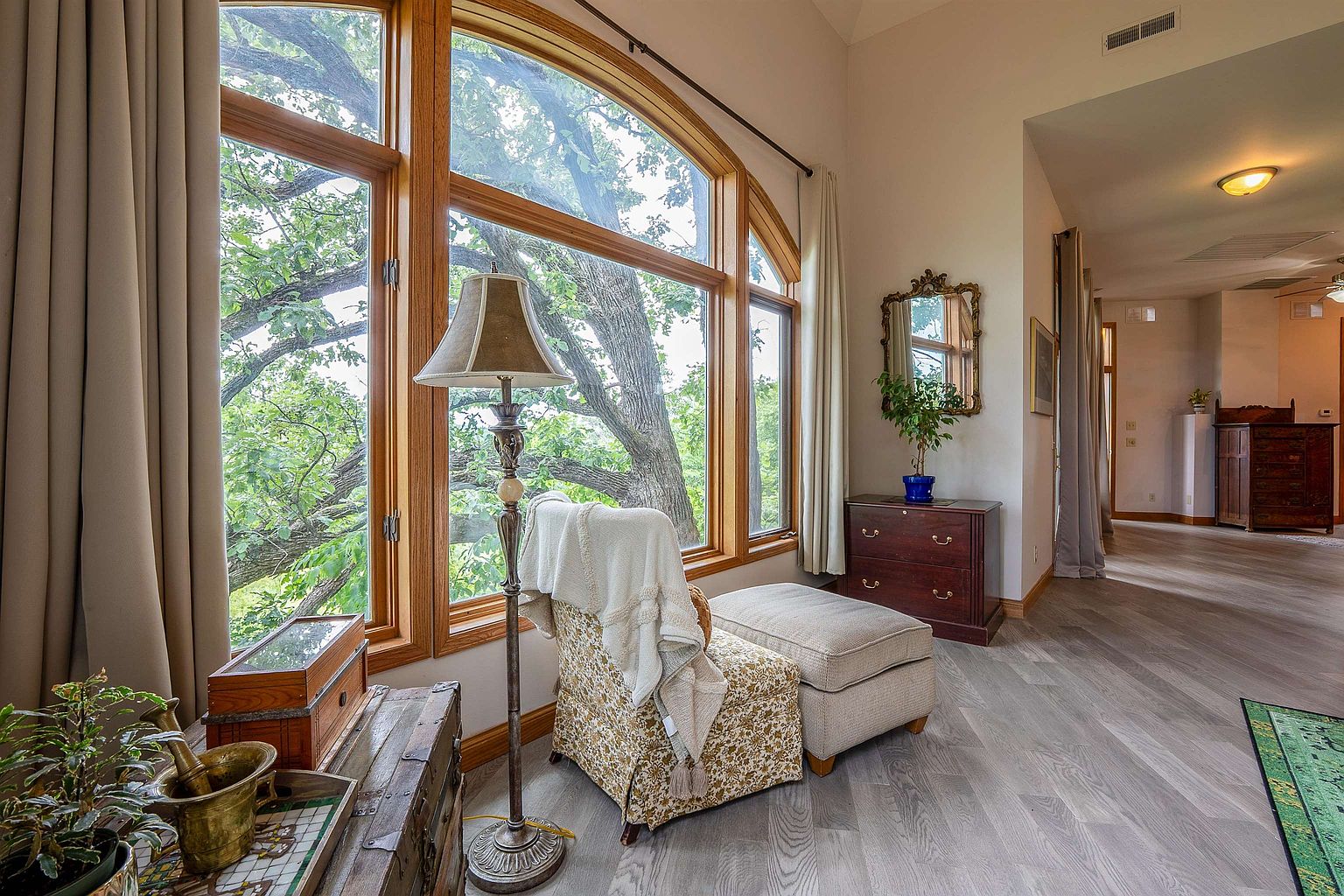 This cozy reading nook features a patterned armchair with a matching ottoman positioned beside a large, multi-pane window that offers a scenic view of mature trees. A tall, ornate floor lamp stands next to the chair, while a dark wood chest of drawers topped with a decorative mirror and a potted plant sits against the wall. The space is finished with light-toned wood-look flooring and flows into a hallway that leads to other areas of the home.