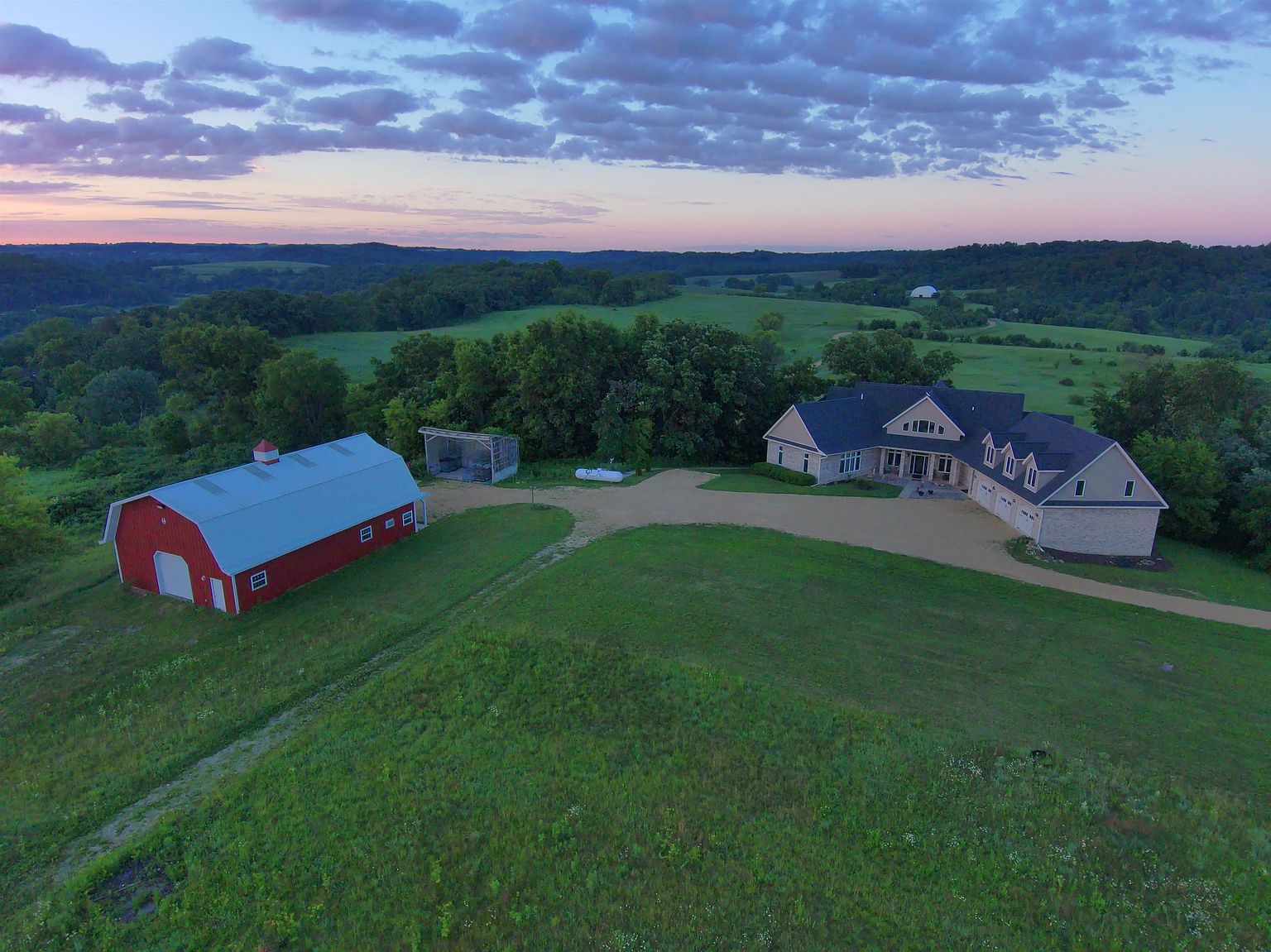 This high-angle aerial view captures a sprawling rural estate at dusk, featuring a large, multi-gabled residence and a classic red barn set against a rolling landscape. A gravel driveway connects the structures, which are surrounded by expansive green fields and mature trees under a soft, colorful sunset sky. The composition emphasizes the property's seclusion and significant acreage, highlighting the harmonious blend of residential and agricultural architecture.