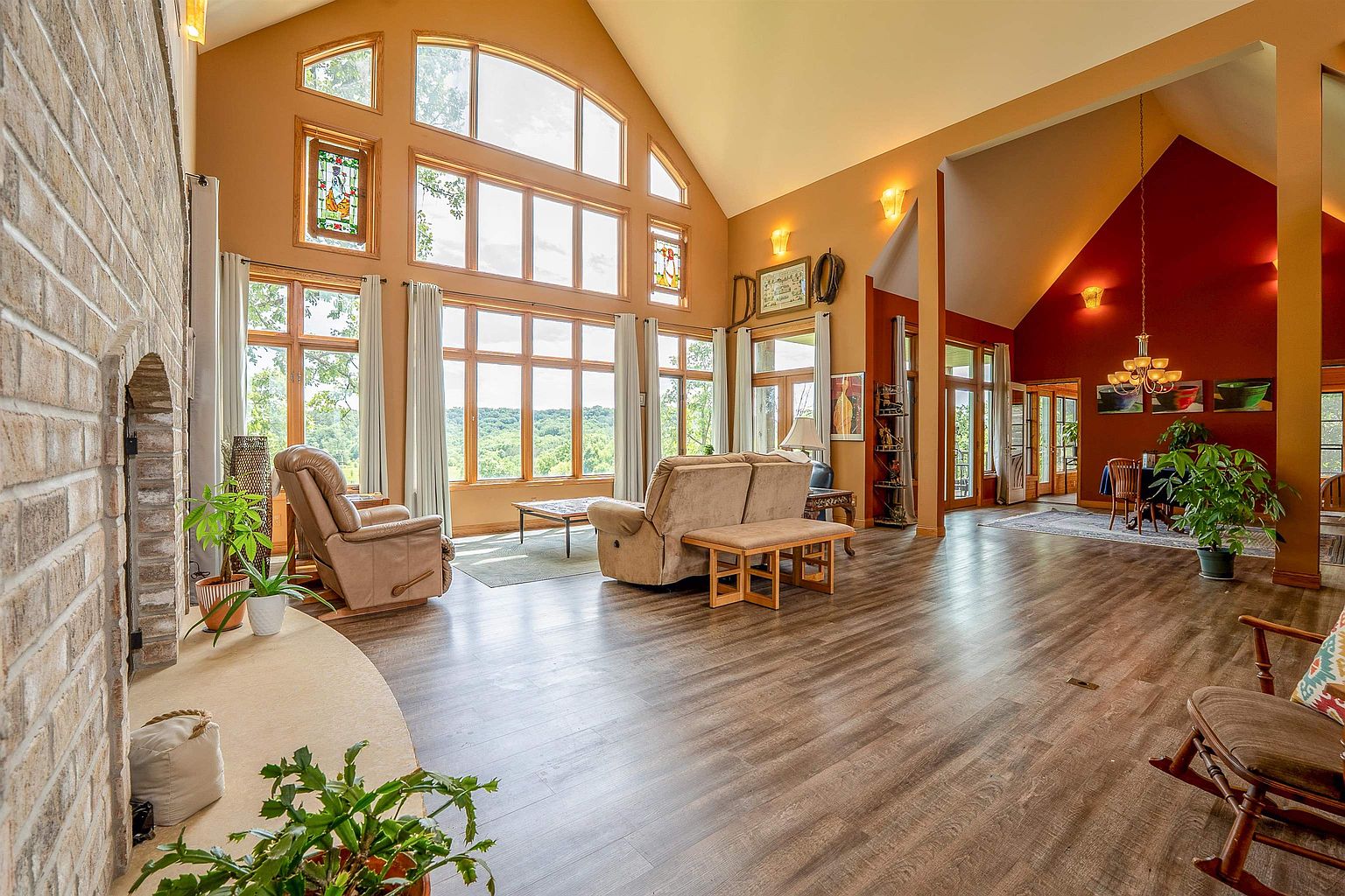 This spacious, open-concept living area features a vaulted ceiling and a prominent stone-clad chimney breast that anchors the room. Large windows provide an expansive view of the outdoors, bathing the space in natural light, while the warm-toned walls and wood-look flooring create an inviting atmosphere. The layout flows seamlessly into a dining area with a bold red accent wall, highlighting the home's airy and connected design.