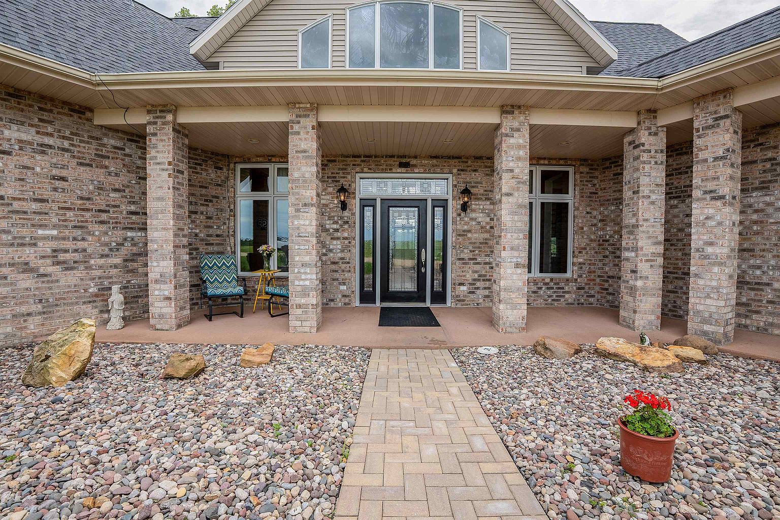 This inviting front entryway features a covered porch supported by substantial brick columns, leading to a central glass-paneled front door. The exterior is finished in a textured brick facade with light-colored siding above, complemented by a stone-paved walkway and a decorative rock garden in the foreground. The perspective is a straight-on, eye-level shot that highlights the symmetrical architecture and welcoming curb appeal of the home.