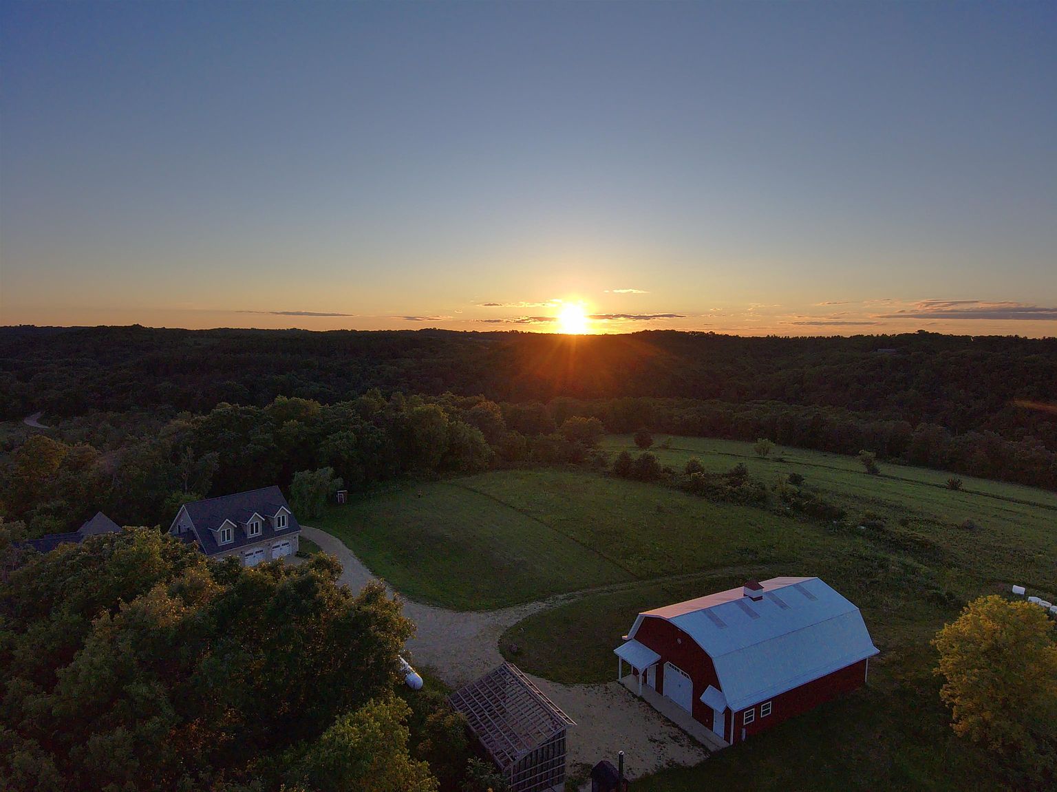 This high-angle aerial view captures a rural property at sunset, showcasing a traditional red barn with a white roof and a separate residential home nestled among mature trees. The expansive landscape features rolling hills and open fields, creating a serene and picturesque setting. The golden light of the setting sun highlights the property's peaceful atmosphere and generous acreage.