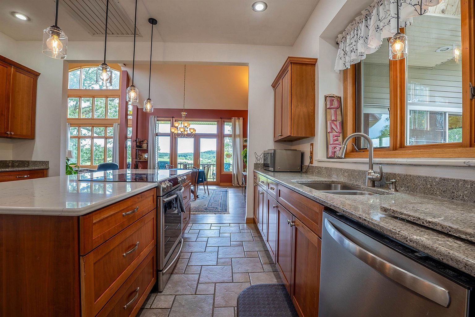 This kitchen features warm-toned wood cabinetry paired with speckled stone countertops and a central island that includes a built-in range. The space is illuminated by three glass pendant lights and flows directly into an adjacent dining area with large windows overlooking a scenic landscape. The overall aesthetic is traditional and inviting, with tile flooring and a functional layout that connects the cooking area to the rest of the home.